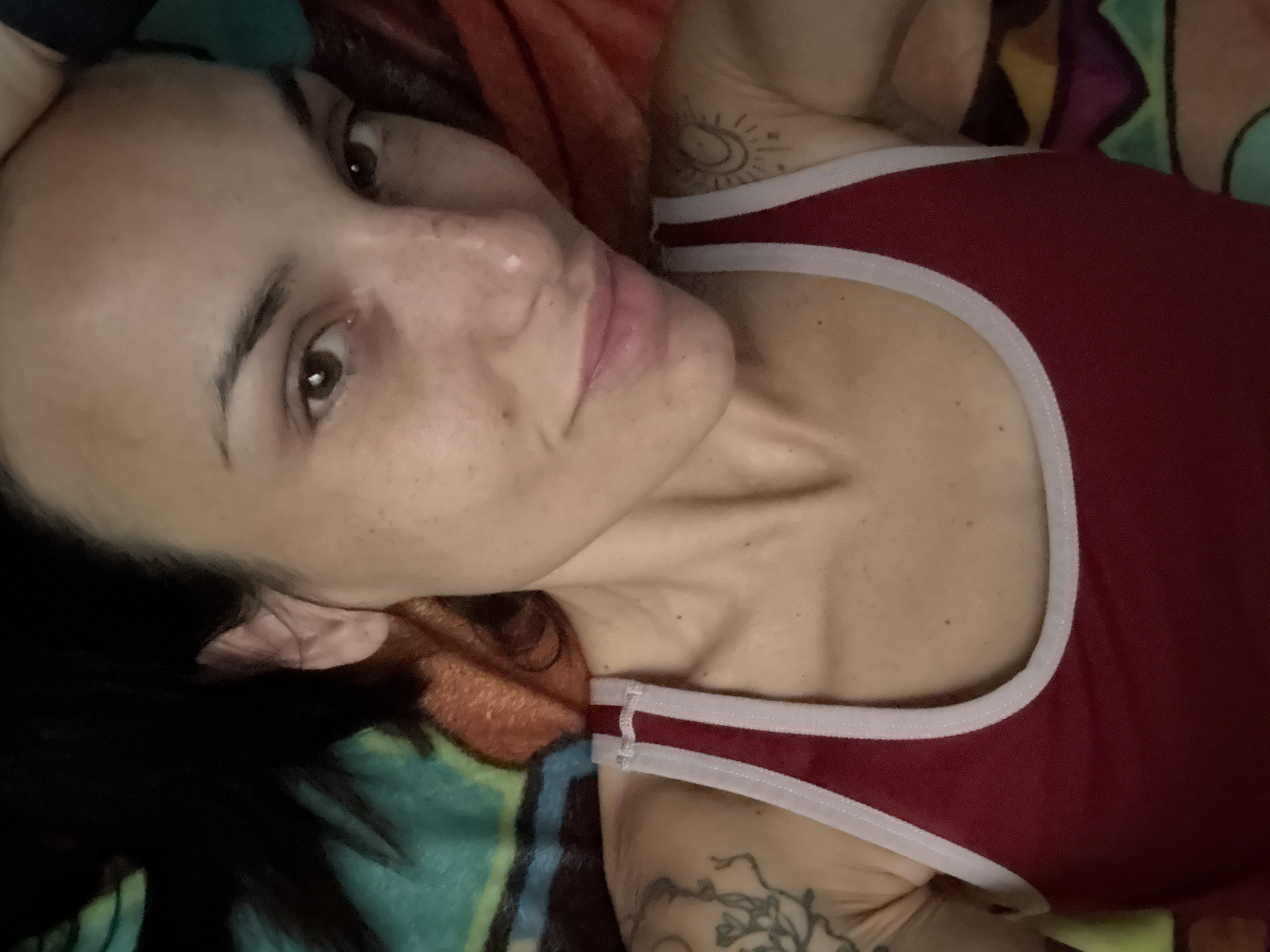 The image is a selfie of a woman with dark hair and brown eyes. She is wearing a red tank top with white trim. She has tattoos on her left arm and shoulder. The background is a colorful blanket. The image is taken from a high angle, looking down at the woman. There are no discernible elements in the image that would indicate the location in the city of Pleven, Bulgaria.