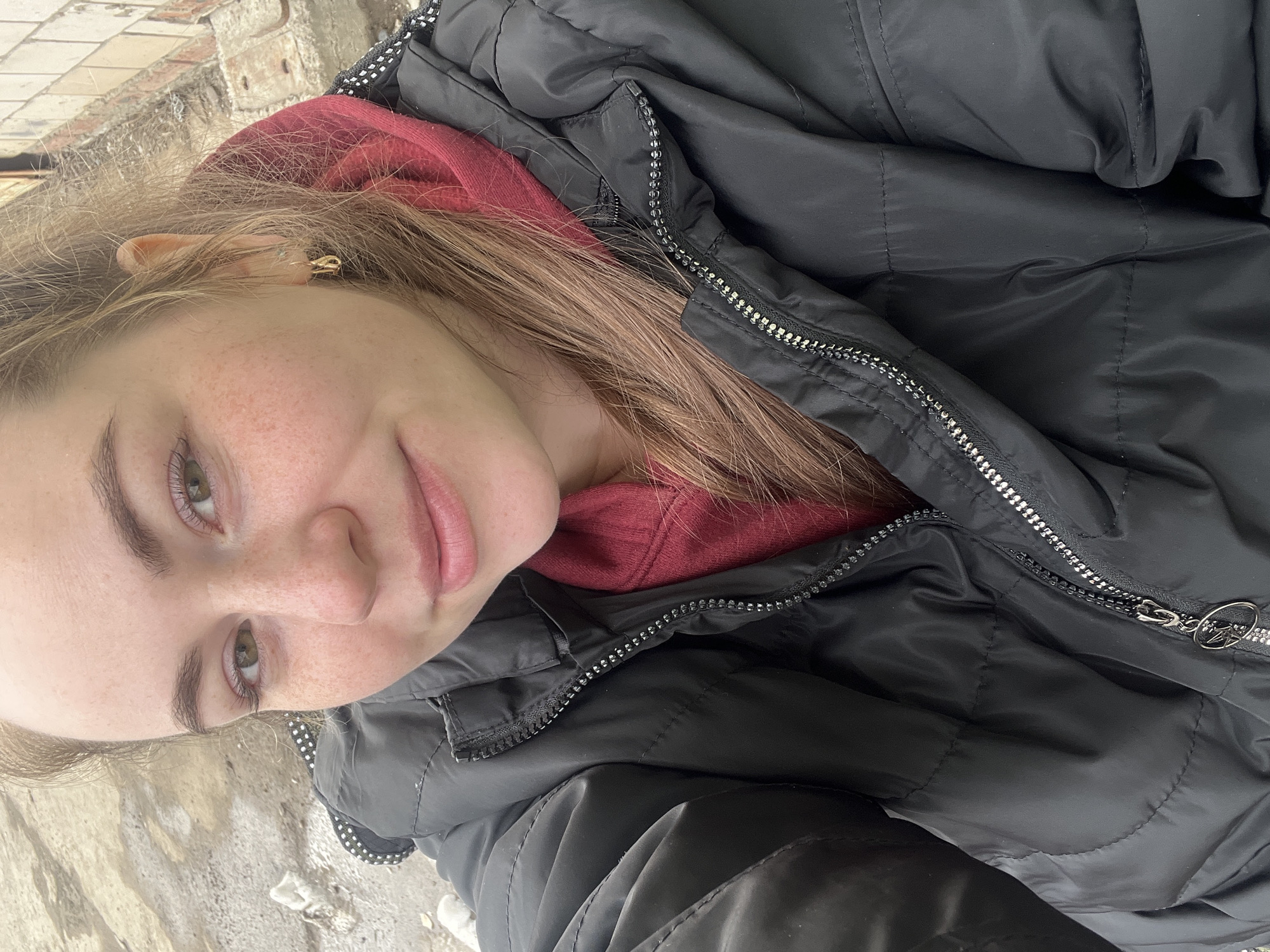 A young woman with light brown hair, freckles, and light eyes is wearing a red hooded sweatshirt and a black puffer jacket. She is outdoors, standing in front of a concrete and brick surface covered with some snow. The image was taken in the city of Chaikovsky, Russia.