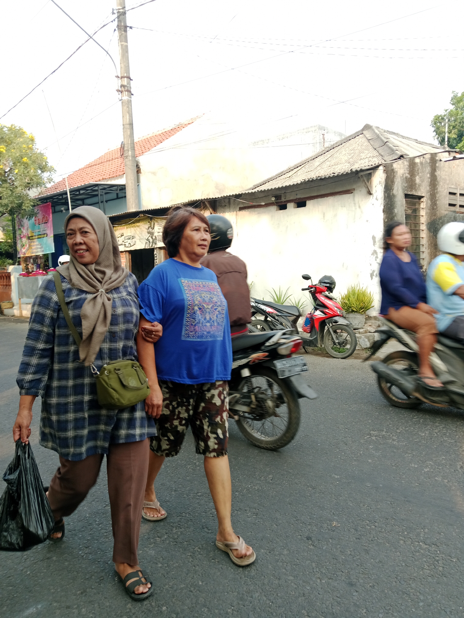 Two women are walking on a street in Kediri City, Indonesia. The woman on the left is wearing a blue and white plaid long-sleeved shirt, brown pants, and a light brown hijab. She is carrying a black plastic bag in her left hand and has a green crossbody bag. The woman on the right is wearing a blue t-shirt with a colorful pattern and camouflage-print shorts. They are walking side-by-side, with the woman on the left's arm linked through the woman on the right's arm. In the background, there are several motorcycles, including a red and black one parked and a black one in motion. A person wearing a brown jacket and helmet is partially visible behind the two women, appearing to be on a motorcycle. Further in the background, there are buildings with corrugated roofs and utility poles with wires. The ground is paved asphalt. The lighting suggests it is daytime.