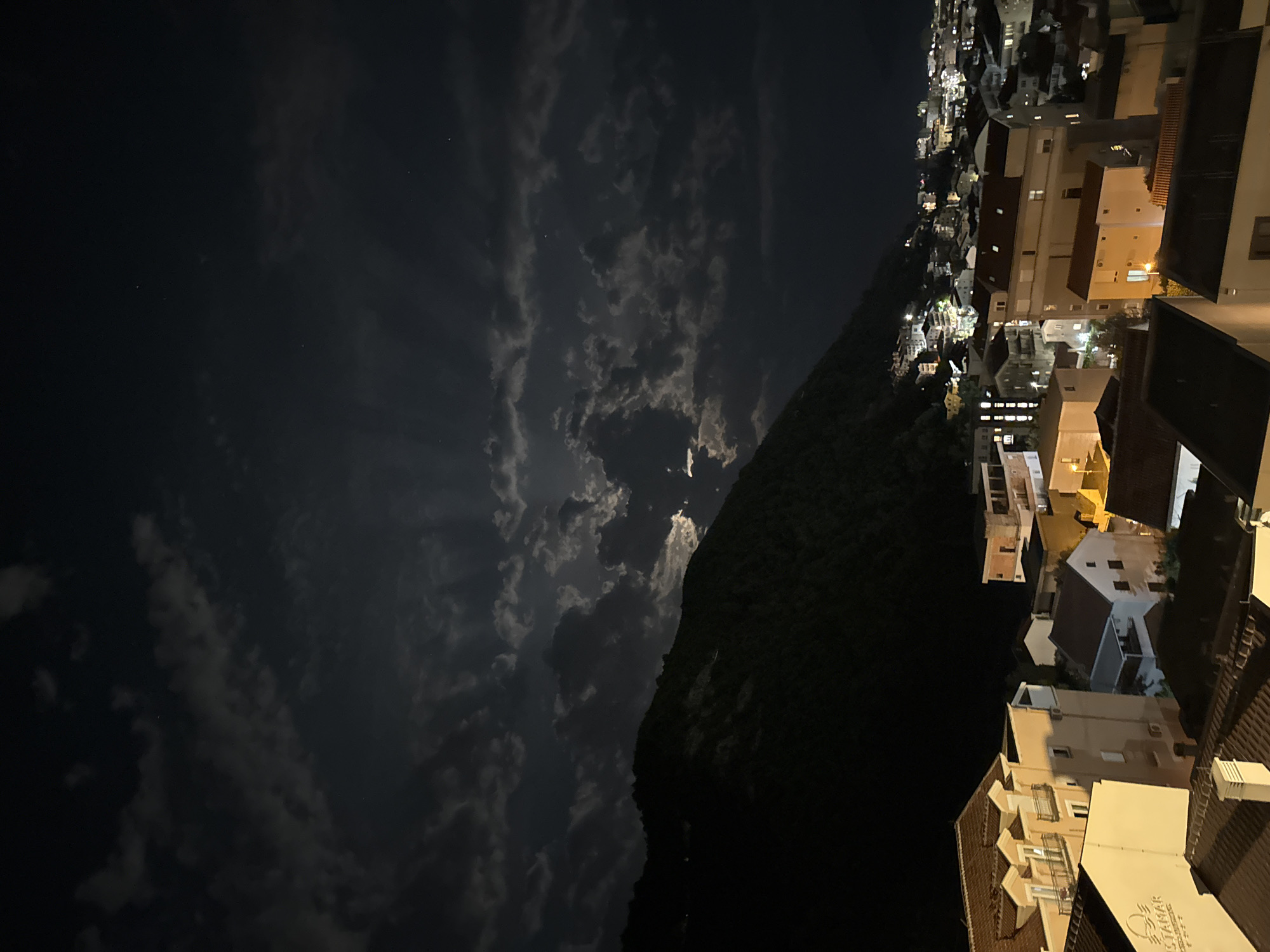 This media file presents a panoramic nighttime view of a town, likely Budva in Montenegro, situated against a formidable, dark mountain. The sky above is a canvas of dark clouds, intermittently illuminated by a visible moon, which casts soft light rays downwards through the cloud cover. Faint stars are discernible in the clearer areas of the night sky.
A large, tree-covered mountain or hill dominates the middle-left portion of the image, appearing as a dark, imposing silhouette. To the right, the town unfolds with a dense arrangement of buildings featuring various architectural styles, including flat and tiled roofs. Numerous windows and streetlights provide artificial illumination, casting a warm glow across the urban landscape and contrasting with the dark natural surroundings. On a building in the foreground towards the bottom right, the text "STAMAR APARTMANI" is clearly visible, suggesting accommodation or a resort. The overall scene conveys a serene and atmospheric coastal night.