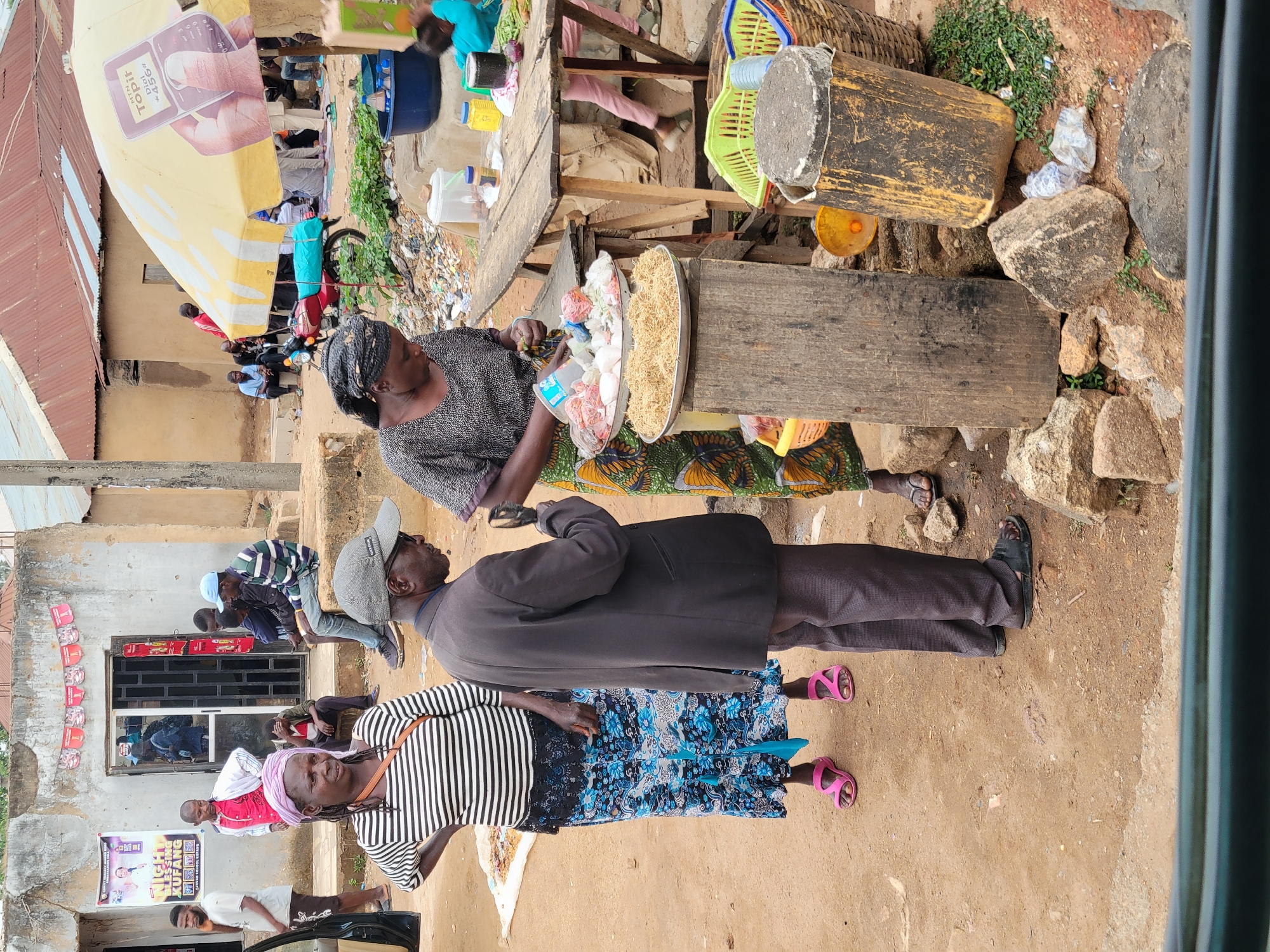 The image depicts a street scene in Jos, Nigeria. A woman is selling food from a table. The food appears to be a variety of small items, possibly snacks or sweets. Two customers stand in front of the table. One is wearing a dark suit and a cap, while the other is wearing a striped shirt and a blue floral skirt. People are gathered near a building in the background. An umbrella with a phone advertisement is partially visible. Debris litters the ground near the food stall. The environment appears to be a busy, commercial area.
