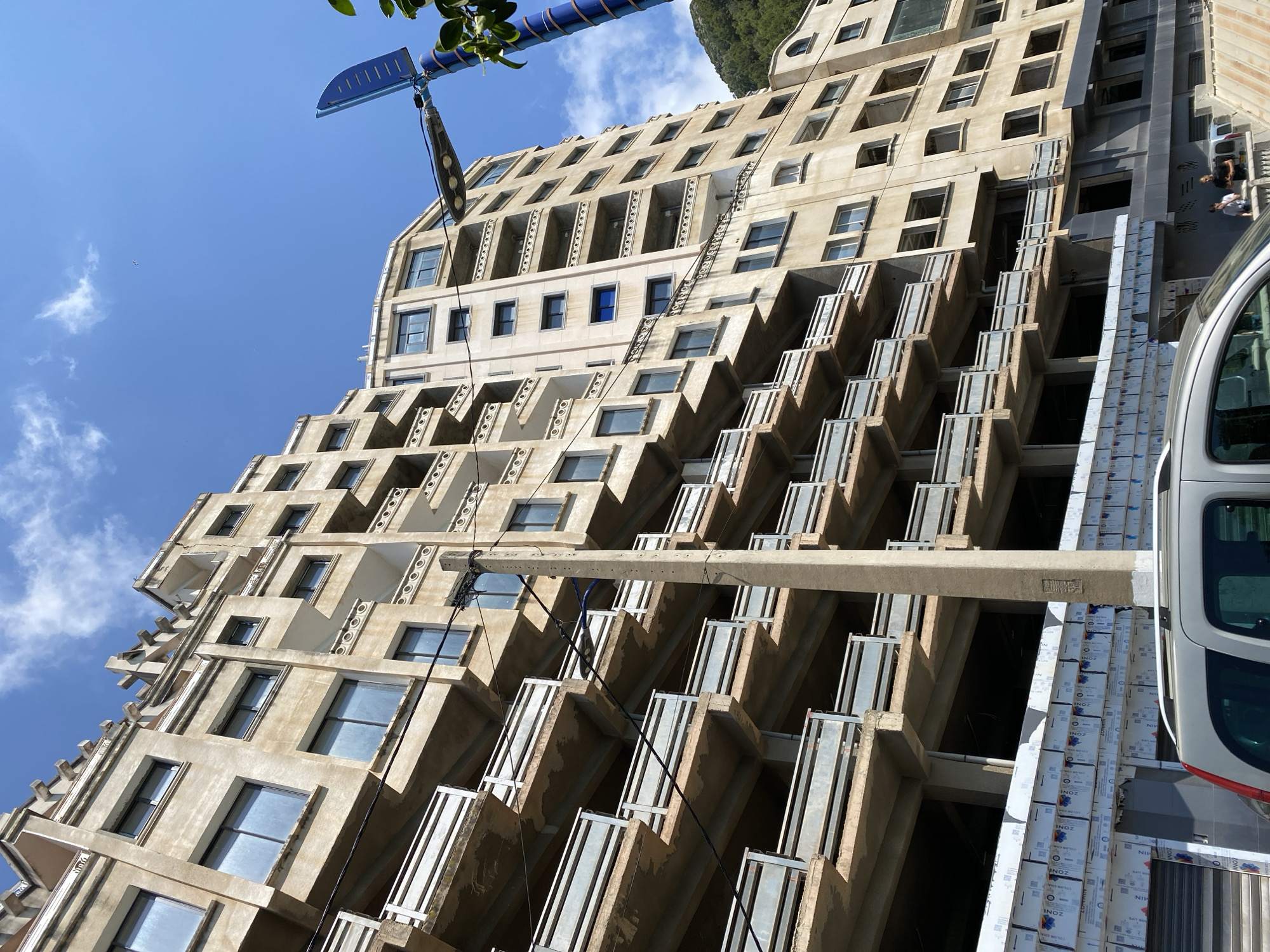 This upward-angled, daytime photograph captures a large, terraced building under construction or renovation in Bejaia, Algeria, against a clear blue sky with sparse clouds. The multi-story structure dominates the frame, featuring numerous rectangular windows and staggered balconies with metal railings, giving it a stepped appearance. Much of the building's concrete framework is exposed, indicating ongoing work.
In the upper left, green foliage is visible, and above the building, part of a blue crane or construction arm extends, with a suspended, elongated object. A small bird flies high in the sky. To the right, a distant mountain or hill is partially visible.
The lower right corner shows the front section of a white van or car, with two individuals – an adult and a child – standing next to it, observing the scene. Stacks of packaged construction materials, possibly facade panels or insulation, are neatly arranged on the ground and along the lower levels of the building. These packages bear text, including repeated instances of "INOZ," along with "ZONA," "NIV," "COLOR CAM," and various numbers like "10000," "9000," and "30000."
The bright sunlight suggests a clear, sunny day, casting distinct shadows on the building's features. The image conveys a sense of active development or transformation within an urban or semi-urban environment.