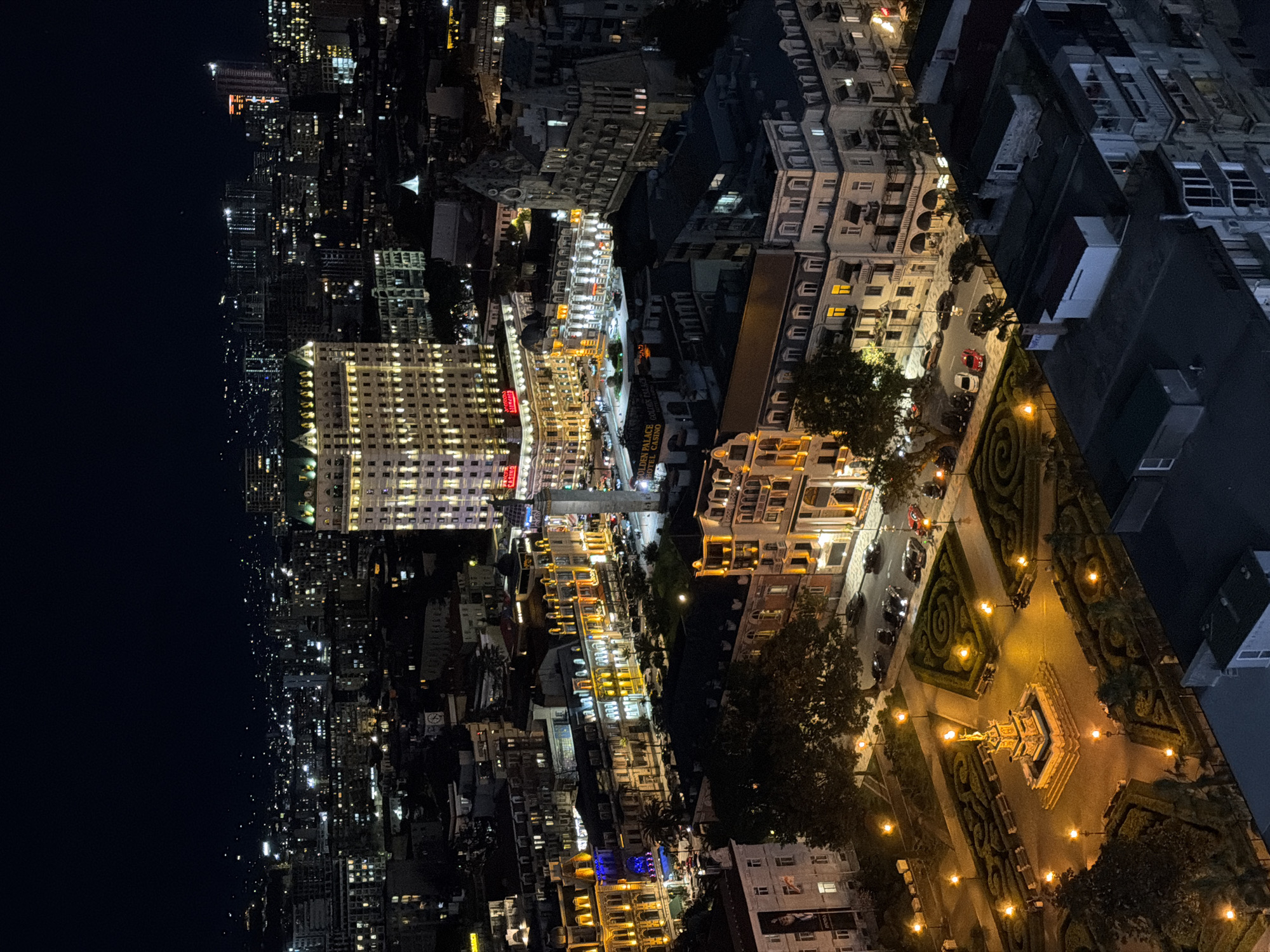 The image shows an aerial night view in Batumi, Georgia. The scene is characterized by a mix of illuminated buildings and infrastructure. On the right side, a dark building can be seen. Adjacent to it is a public park decorated with a fountain and spiral patterns. A line of cars is parked along the road on one side of the park. The central area is composed of bright commercial buildings with some signage visible, along with a visible road. On the far left, a long street with many tall apartment buildings extends into darkness.