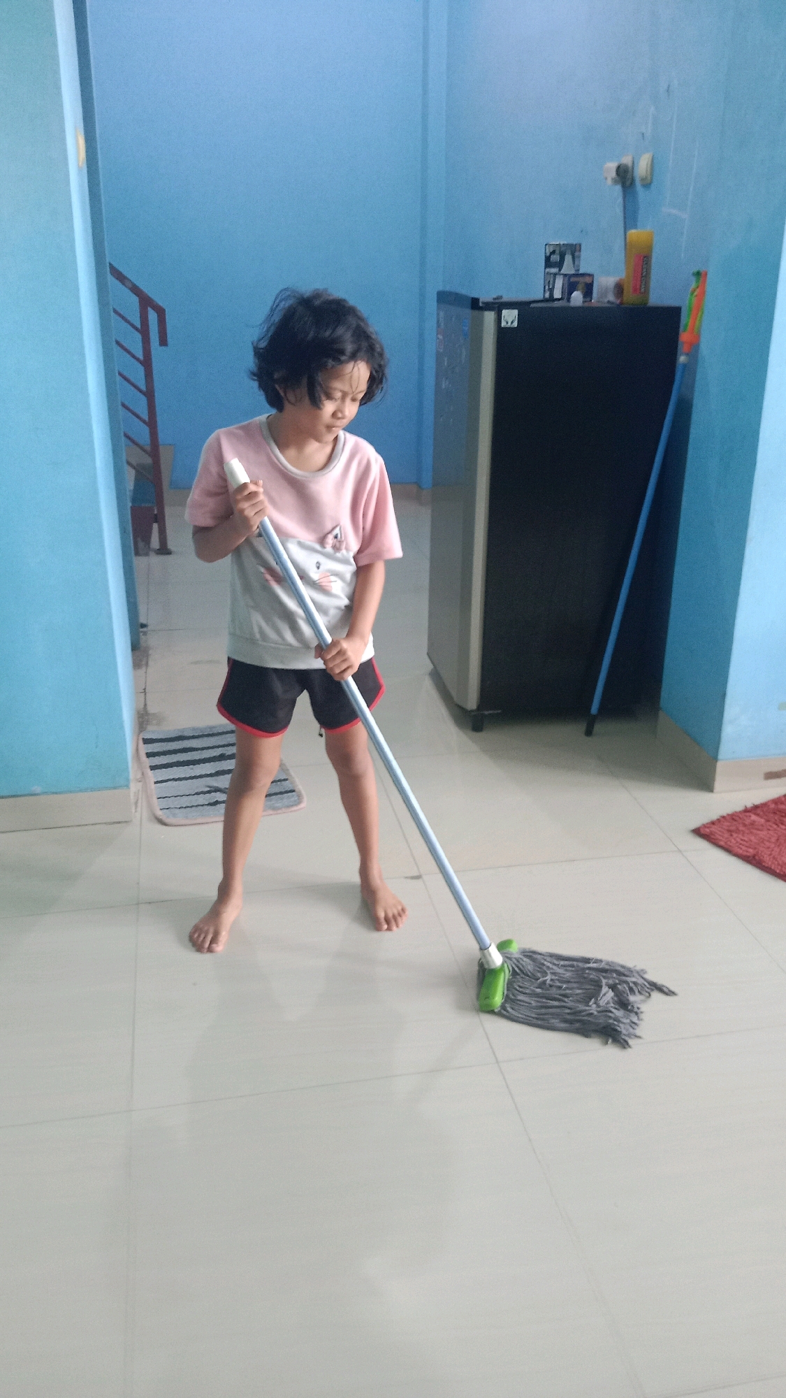 In a room with light blue walls and a tiled floor, a young individual with dark, messy hair is mopping the floor. They are wearing a pink and gray t-shirt and black shorts with red trim. The individual is barefoot and is holding a mop with a green mop head and a light blue handle. To the right of the individual, a refrigerator is visible, and behind it, a broom with an orange handle is leaning against the wall. A staircase with metal railings is also present in the background. The scene suggests a residential interior in Jalenjaya, Indonesia.
