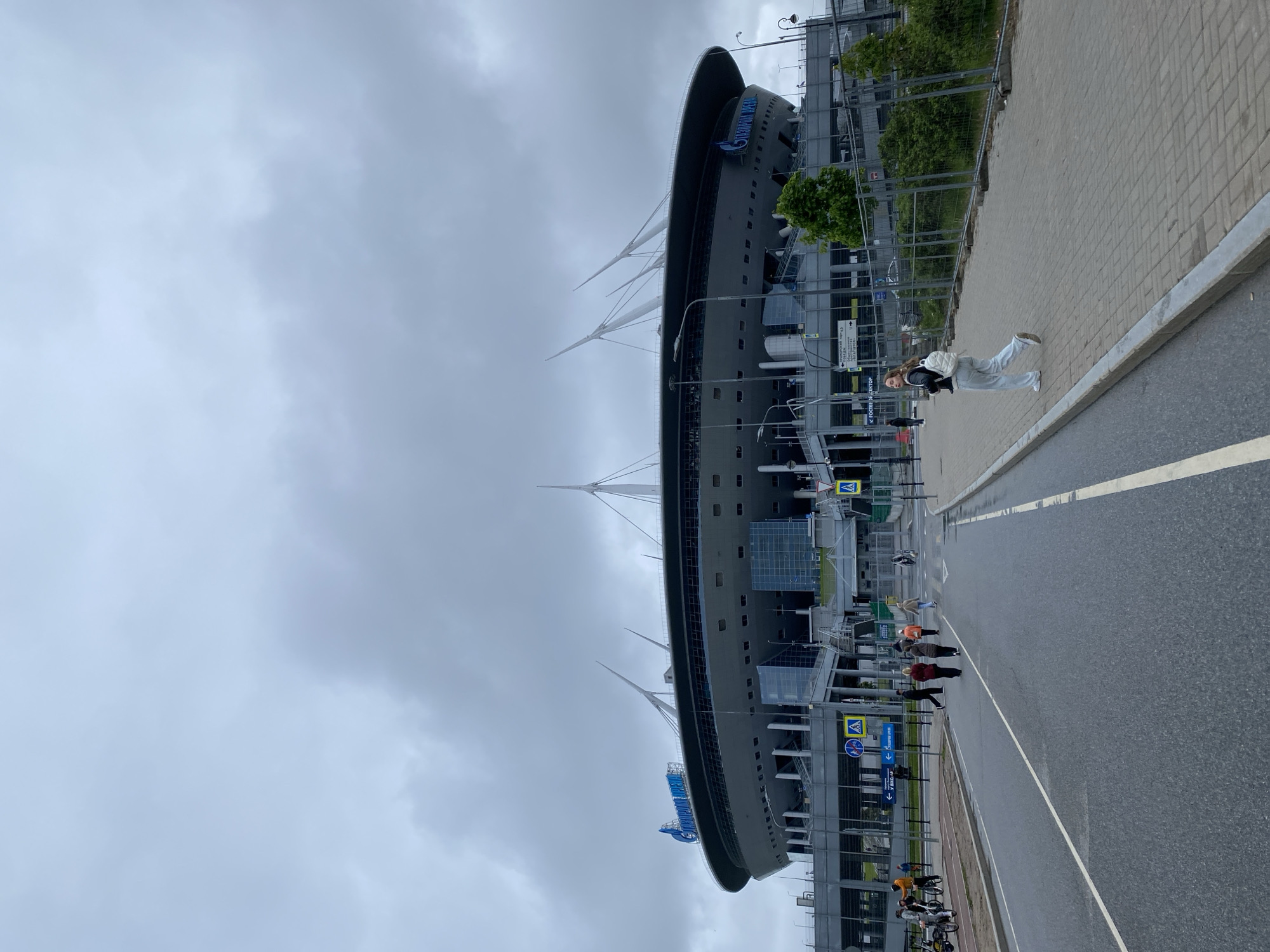 The image shows the Gazprom Arena in Saint Petersburg, Russia, under a cloudy sky. A woman in light-colored clothing is standing on the sidewalk next to the stadium, and a group of people are walking nearby. Bicycles are parked along the fence separating the sidewalk from the stadium. The street is wet, with a white lane marker visible.