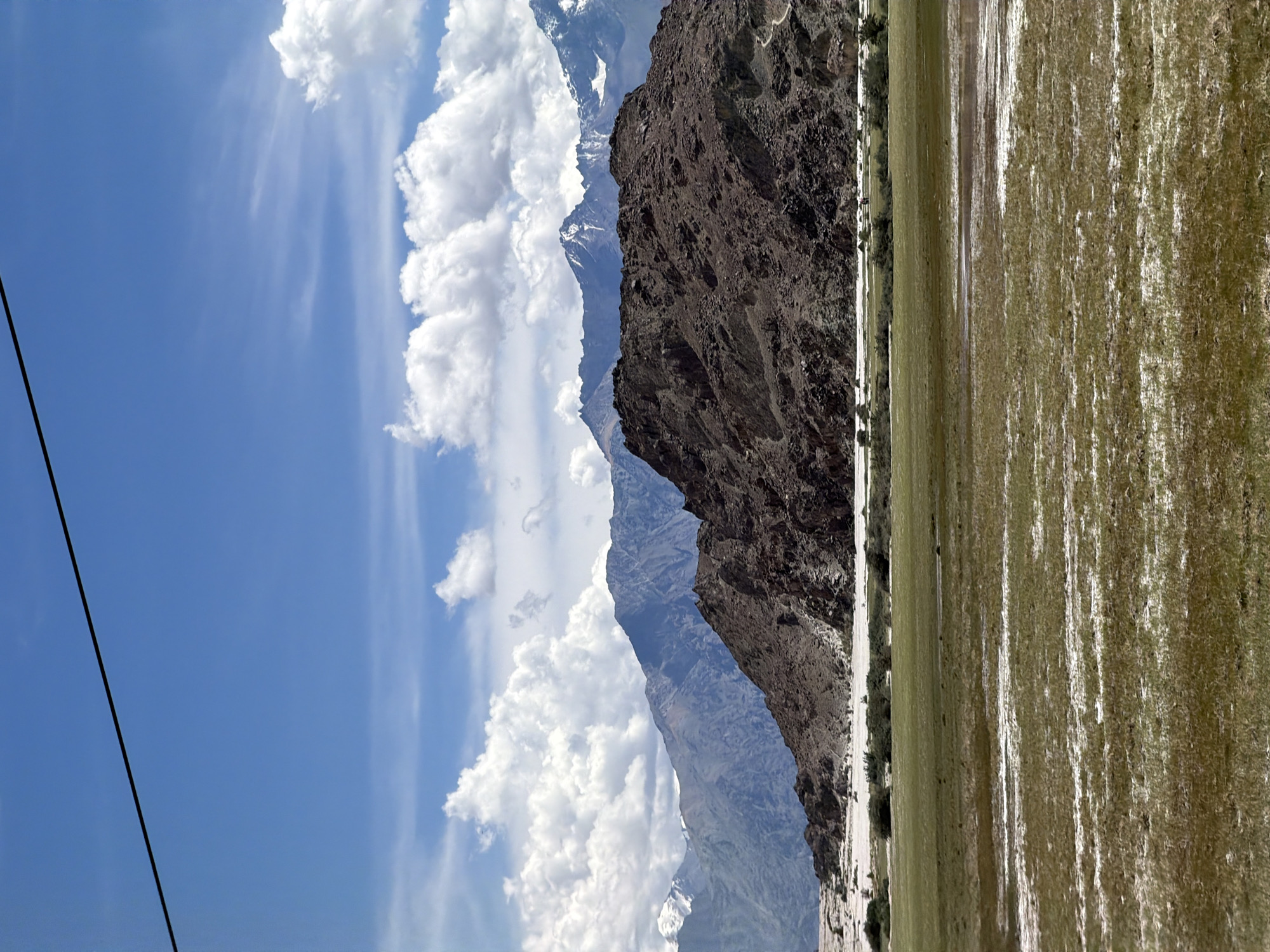 A landscape photograph depicts a vast, mountainous region under a bright blue sky dotted with white, fluffy clouds. In the foreground, a grassy field with patches of damp earth or shallow water stretches across the frame. Further back, a range of mountains rises, with a prominent, dark, rocky hill in the mid-ground. The peaks of the furthest mountains are snow-capped. The scene is devoid of any human presence or structures, conveying a sense of remote, natural beauty. The image is captured during daylight hours.