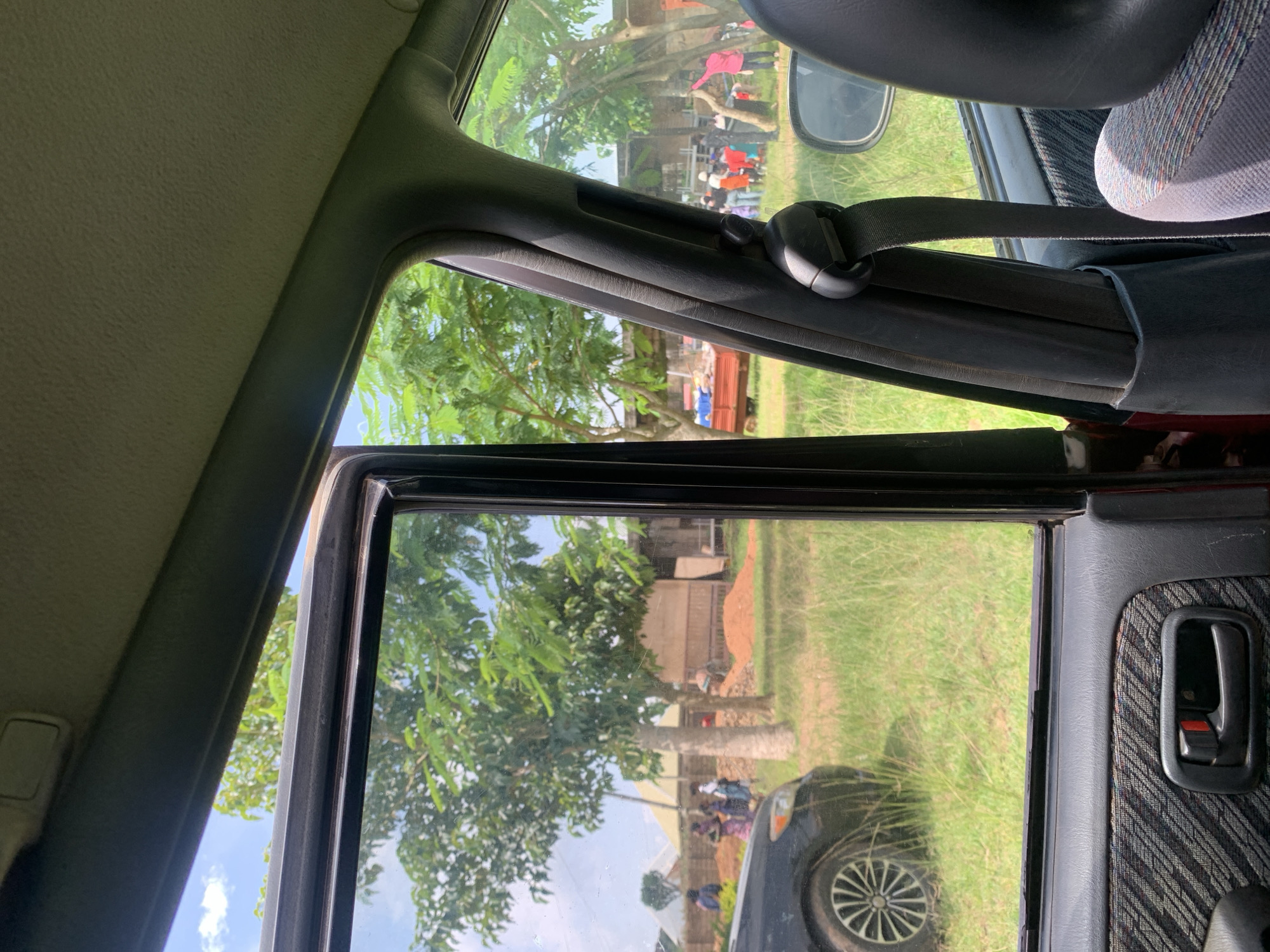 An outdoor scene is viewed from inside a car. Through the open car window, a grassy area is visible with trees. In the background, several people are gathered near a building and a red trailer. There is also a dark-colored car in the foreground, partially visible on the left side of the frame. The sky is partly cloudy. The interior of the car shows part of a seat, a seatbelt, and the door panel with a handle. The location is described as Unknown, Unknown.