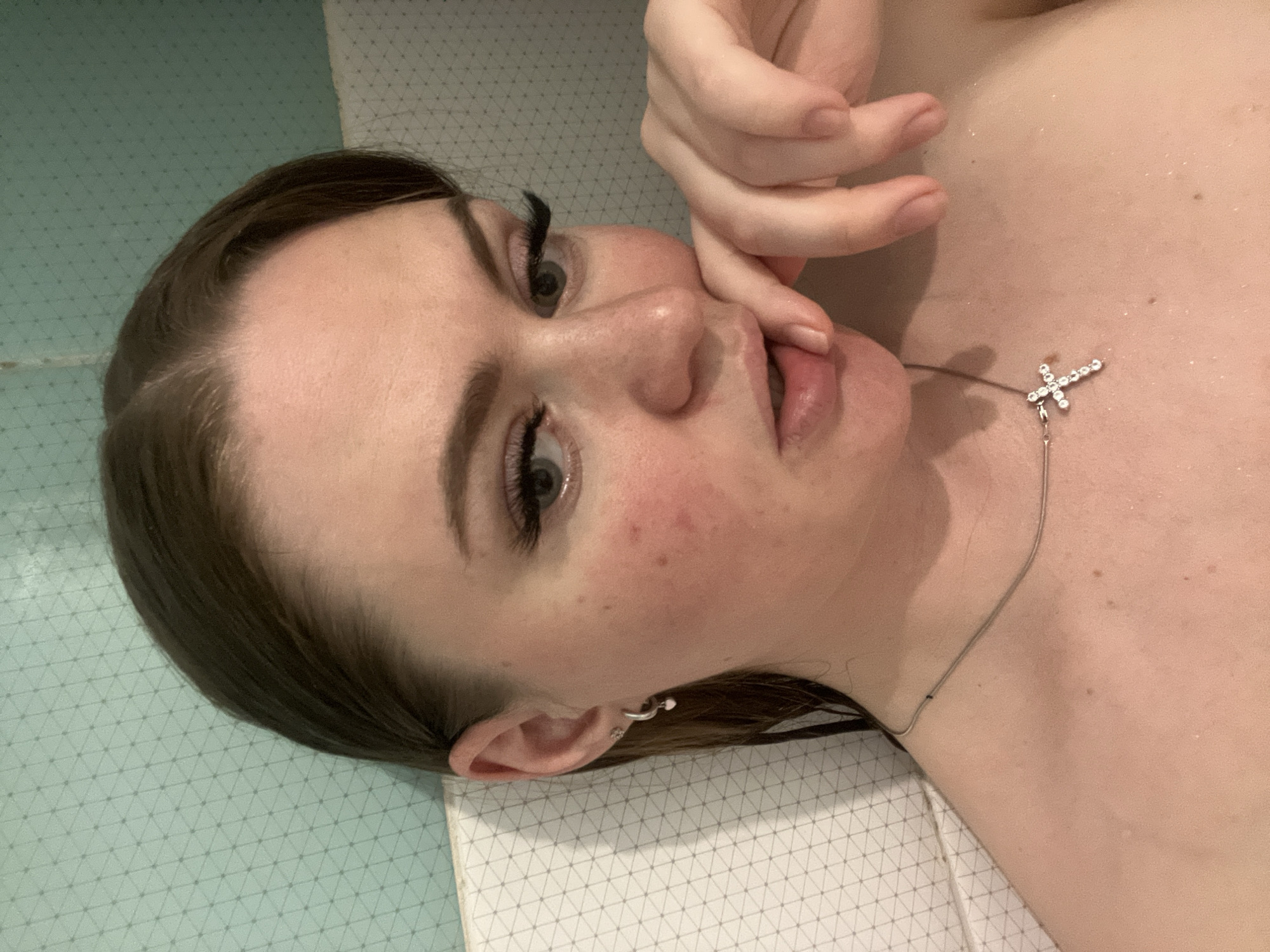 A close-up, portrait-oriented photograph depicts a young woman lying on a surface with a geometric pattern, possibly tiles, as suggested by their color and arrangement. The woman's face is the central focus, and her features are prominent. She has fair skin, dark eyebrows, long dark eyelashes, and appears to be wearing minimal makeup. Her brown hair is styled back.
She is wearing a silver necklace with a small cross pendant. Her right hand is raised to her face, with her index finger touching her bottom lip. The background is out of focus. No further details about the location are discernible from the image.
