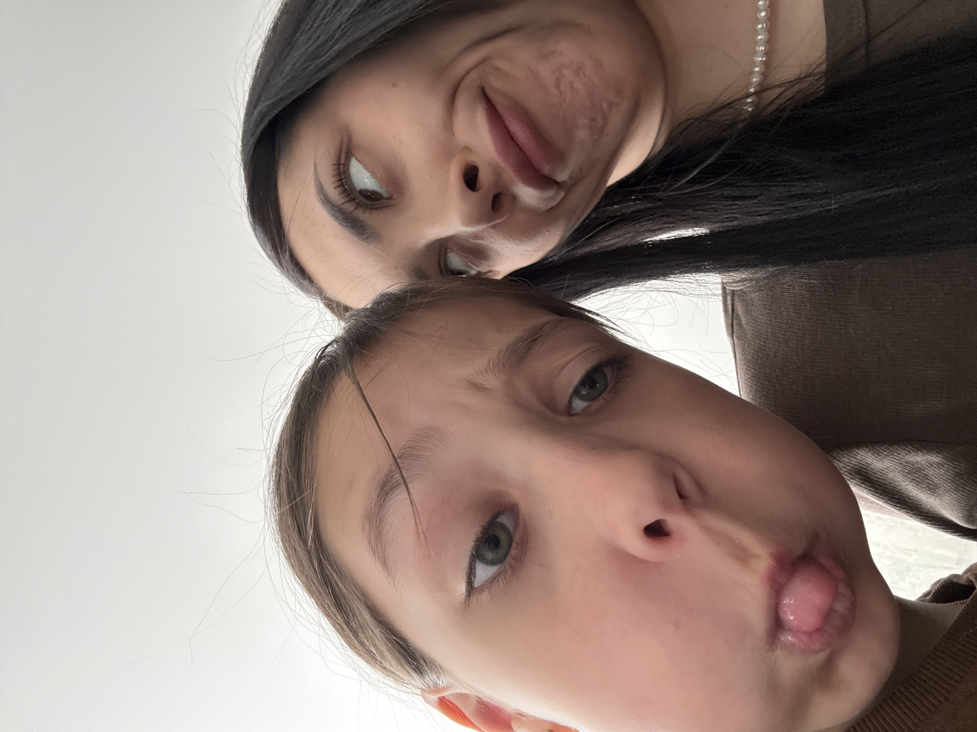 Two individuals are closely positioned, appearing to be taking a selfie. The upper individual has dark hair and is wearing a pearl necklace. The lower individual is younger, with light hair and a visible tongue. The background is mostly a plain white color. The image is angled, presenting an unusual perspective. There is no information to suggest the location is Votkinsk, Russia.