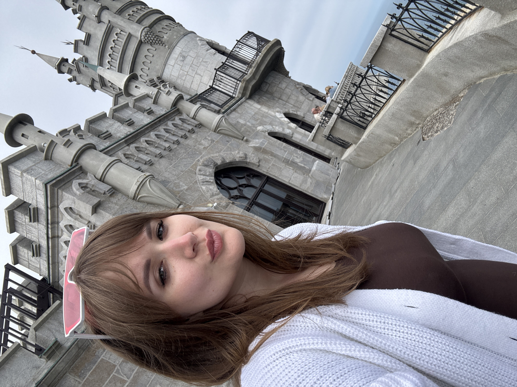 A young woman with brown hair, wearing a brown top and a white cardigan, is posing for a selfie. She is standing next to a grey stone building with turrets and balconies, reminiscent of a castle. In the background, other people are visible on a balcony. The location is the Swallow's Nest castle, in the Гаспринский поселковый совет, Ukraine.