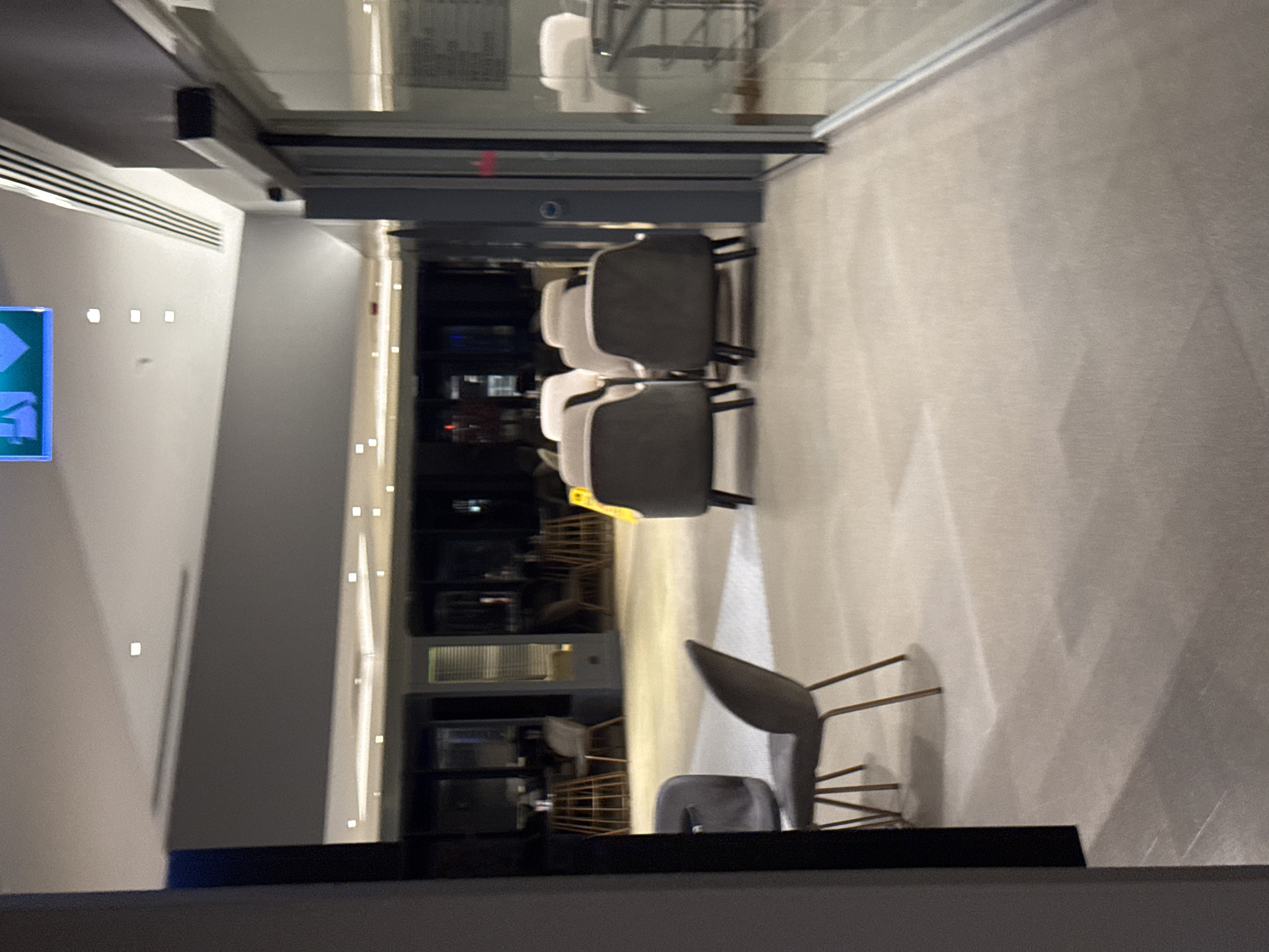 The image captures an interior scene, likely a lounge area or a waiting room, with a focus on seating arrangements and architectural elements. The scene is well-lit, with recessed lighting fixtures visible in the ceiling. An illuminated green exit sign is prominently displayed overhead, indicating a possible emergency exit.
In the foreground, several chairs are arranged. Two dark-colored chairs with slender legs are positioned in the lower left and center of the frame. To their right, a row of lighter-colored chairs forms a seating cluster. Further into the background, more seating is visible, including chairs and small tables, suggesting a functional space for relaxation or waiting.
The environment appears modern and clean, with neutral-toned walls and flooring. A large glass-enclosed area, possibly a shop or another section of the building, is visible on the right side of the image, reflecting the interior. The overall impression is one of a contemporary, organized indoor space, devoid of any individuals in the immediate frame. There are no discernible elements that link this image specifically to the city of Aksu, Turkey.