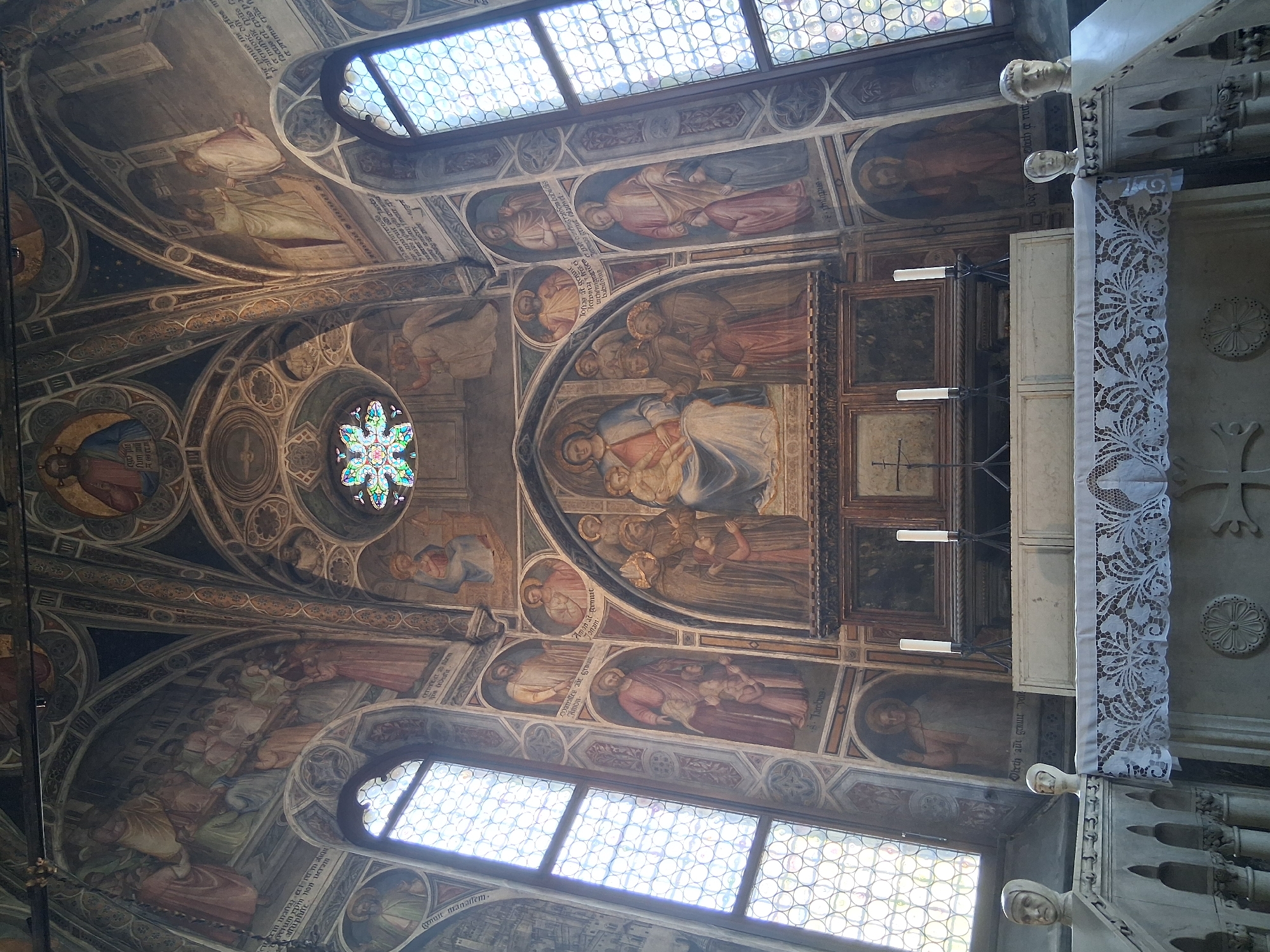 The image shows the interior of the Scrovegni Chapel in Padua, Italy, specifically the altar area. The walls and ceiling are extensively decorated with frescoes. In the center, above the altar, is a large fresco depicting the Virgin Mary holding the infant Jesus, surrounded by saints and other figures. To the left and right of this central fresco are tall, arched windows with intricate stained glass.
The altar itself is draped with a white lace cloth. On the altar are two tall white candles in black holders and a crucifix. The altar is set against a backdrop of more frescoes and architectural elements. In the foreground, there are stone sculptures of heads on either side of a balustrade. The overall impression is of a richly adorned and historic religious space.