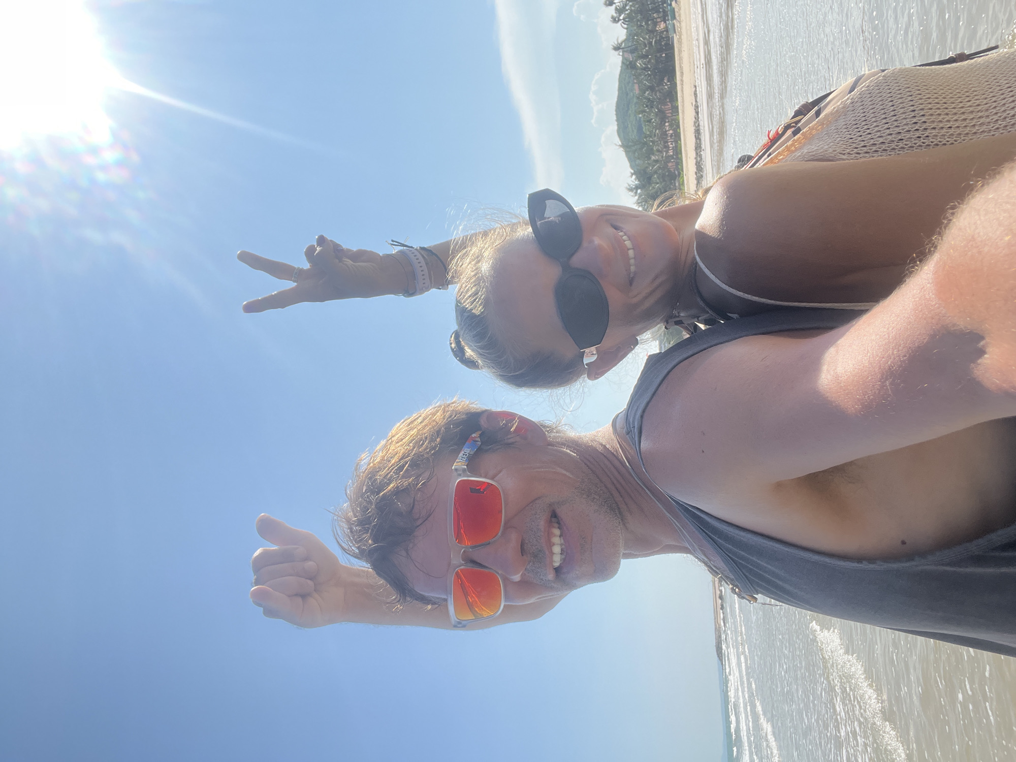 A man and a woman stand back to back on a beach in Phu Thuy Ward, Vietnam, with the ocean and sky in the background. The man, on the left, wears a grey tank top and sunglasses with orange lenses. His left arm is raised with his fist closed in a thumbs-up gesture. The woman, on the right, wears a light-colored crocheted top and dark sunglasses. Her right arm is raised with her index and middle fingers extended in a peace sign. Both individuals are smiling. The sun is bright, causing lens flare in the upper right corner of the image. Palm trees and buildings can be seen in the distance along the coastline.