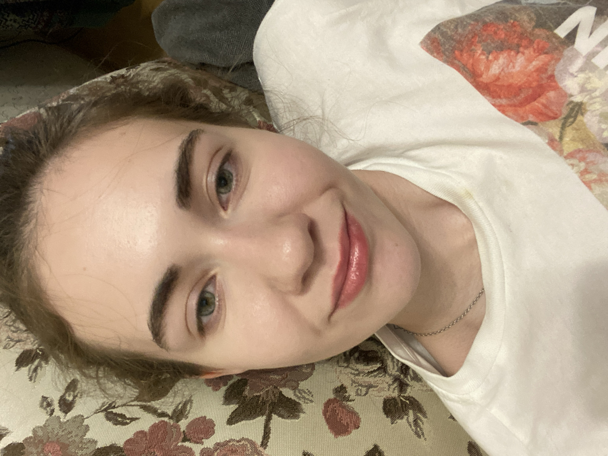 The image shows a young woman lying on a floral-patterned surface, possibly a sofa or bed, in Zavodoukovsk, Russia. She is wearing a white t-shirt with a floral print and a silver chain necklace. Her head is tilted, and she is looking directly at the camera with a slight smile. Her eyes are green and her lips are colored with a pinkish-pink lipstick. The lighting suggests it is daytime, but there are no clear indicators of the weather or the specific time of day. The background is out of focus, but appears to include a dark grey fabric, possibly a blanket or another piece of furniture. The overall impression is casual and relaxed, with the woman appearing comfortable and at ease. There is no visible text in the image.