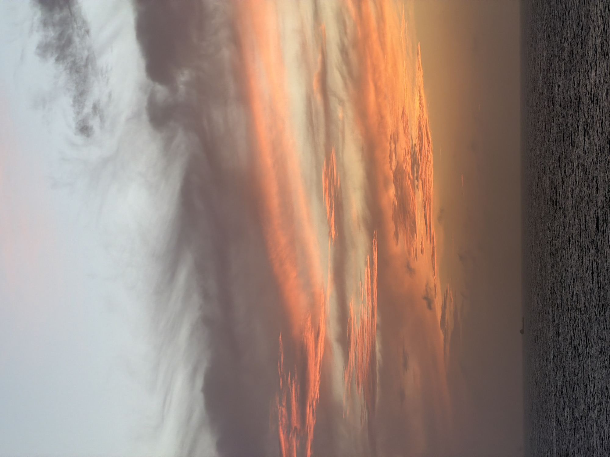 The image shows a sunset over the ocean. The sky is filled with clouds, which are illuminated in shades of orange and pink. The ocean is dark and choppy. A sailboat is visible on the horizon. The location is not identifiable from the image.