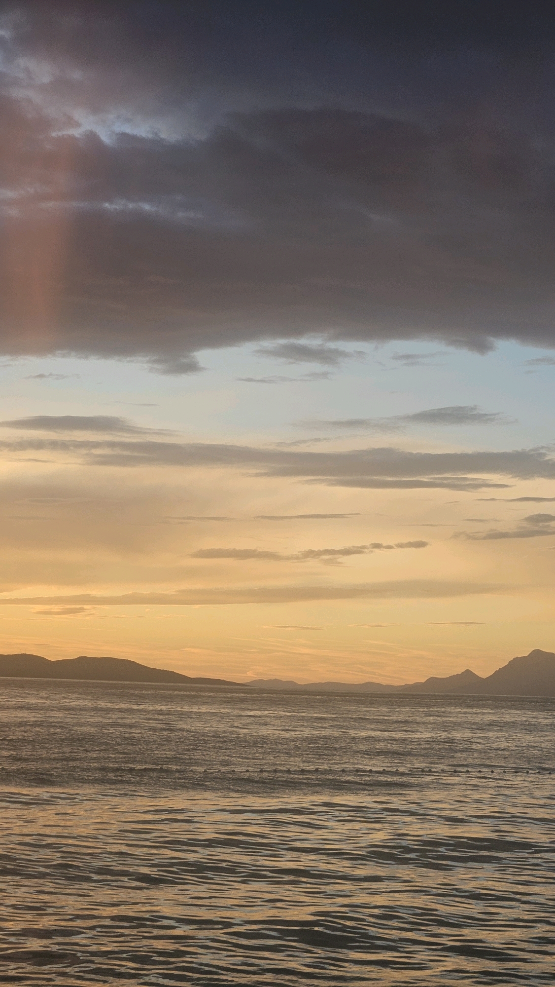The image depicts a seascape at dusk or dawn, likely near Općina Tučepi, Croatia. The sky is a gradient of dark, moody clouds at the top transitioning to lighter shades of blue and then to a warm, golden orange hue near the horizon. The sea is calm with gentle ripples, reflecting the colors of the sky. In the distance, silhouetted landmasses, possibly mountains or islands, form the horizon line. A line of small, spherical buoys is visible on the water's surface, marking a boundary or swimming area. The overall impression is one of tranquility and natural beauty, characteristic of the Adriatic coast.