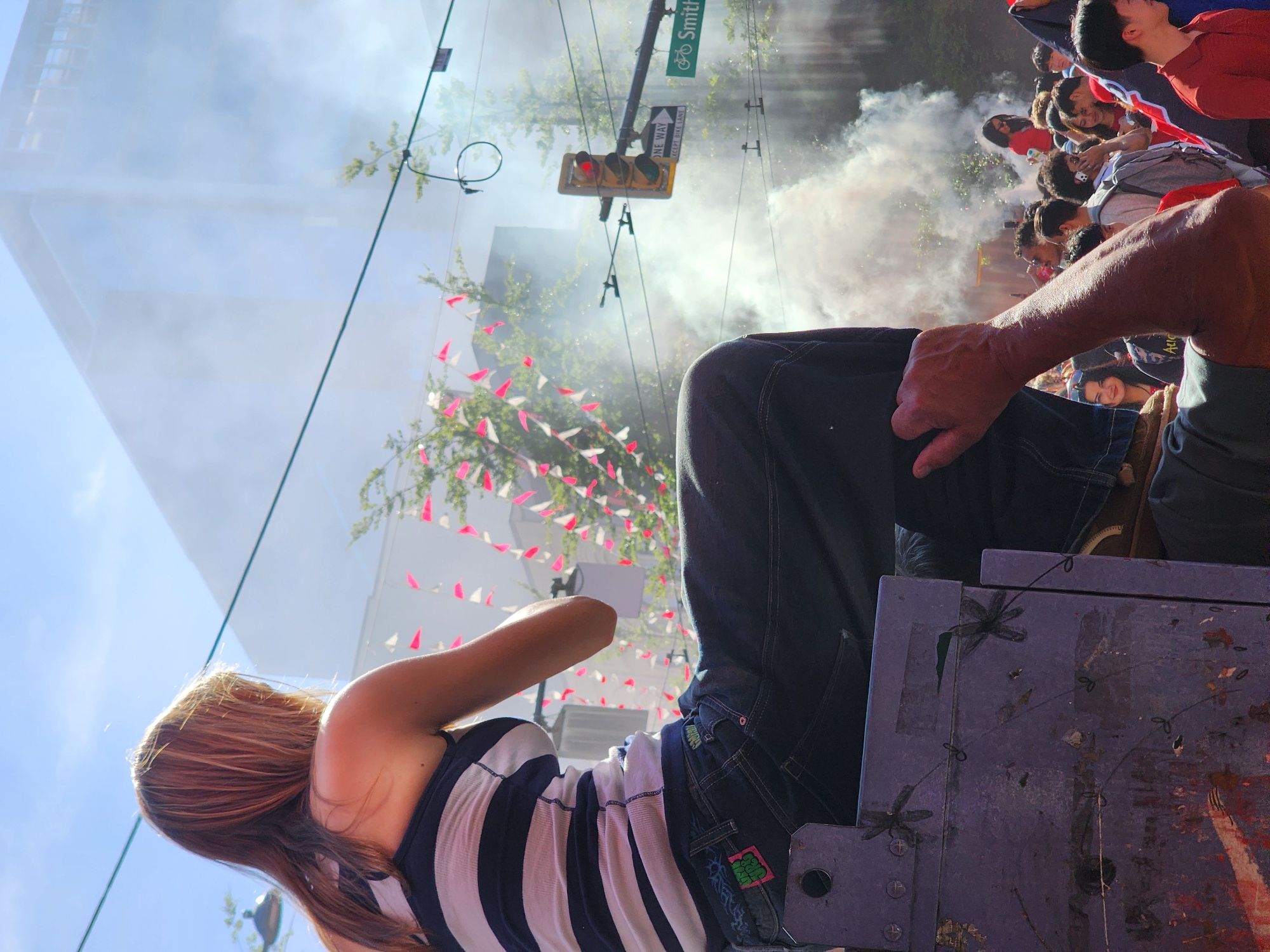 A woman with long blonde hair sits facing away from the camera on a purple object in the foreground of a street scene. She wears a navy and white striped tank top and dark denim jeans, with a green and pink patch on her left back pocket. Behind her, a crowd of people gathers, some holding up cell phones. Smoke billows in the air, obscuring the background buildings and trees. A traffic light with a red signal hangs above the crowd. A green street sign reading "Smith" is visible on the right. The sky is blue with scattered clouds. The overall impression is a vibrant, possibly celebratory, outdoor event.