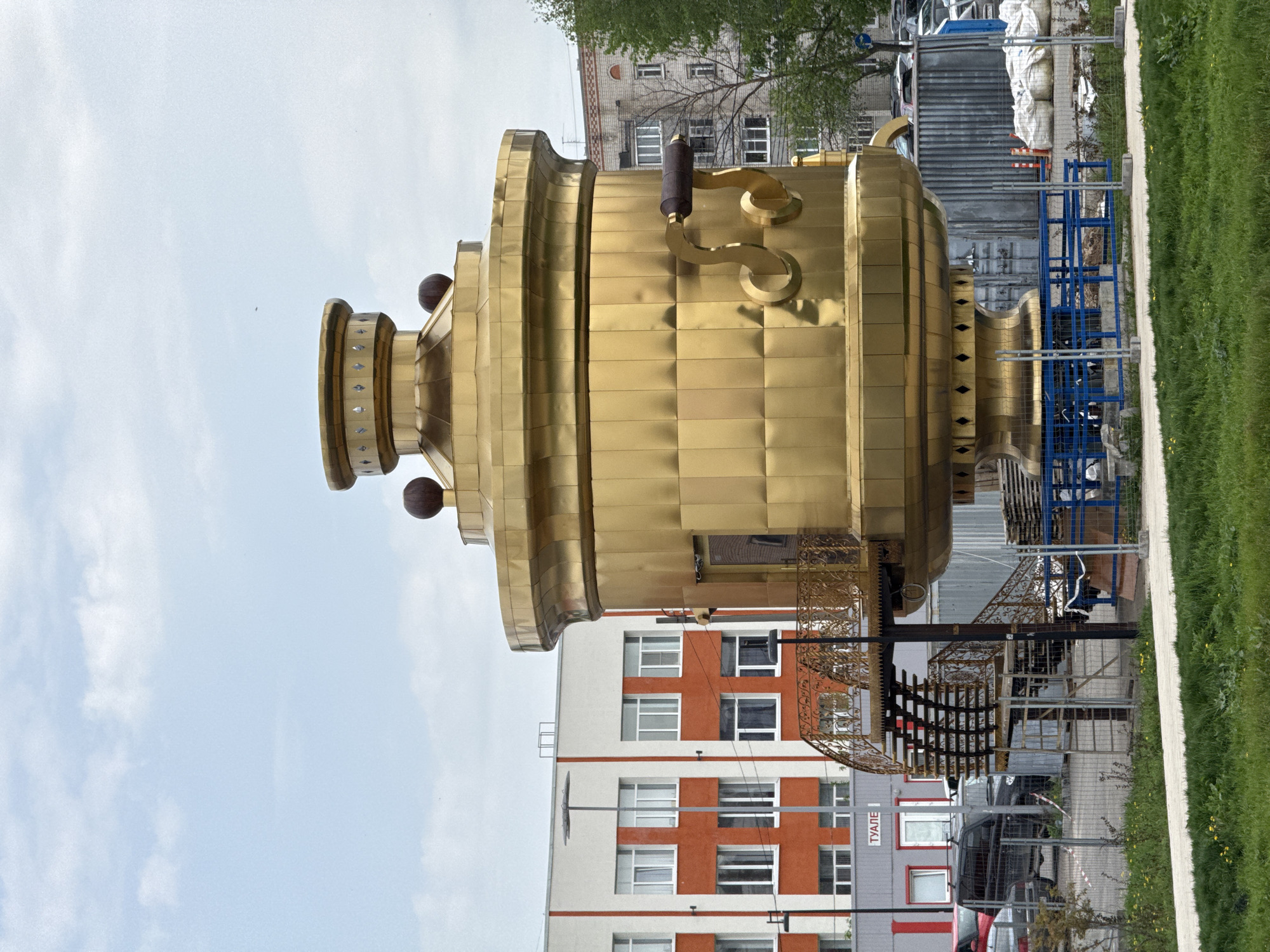 The image shows a large, golden monument of a samovar (a traditional Russian tea urn) located in the city of Bor, Russia. The samovar is ornate with a cylindrical body composed of rectangular, golden panels, a top section with decorative spheres, and a faucet. Underneath it is a dark, wrought-iron structure. In the background is a modern building with a white and orange façade. Next to the samovar, there are construction materials and equipment, including blue scaffolding and metal containers. A strip of green grass runs along the right edge of the image. The sky is overcast.