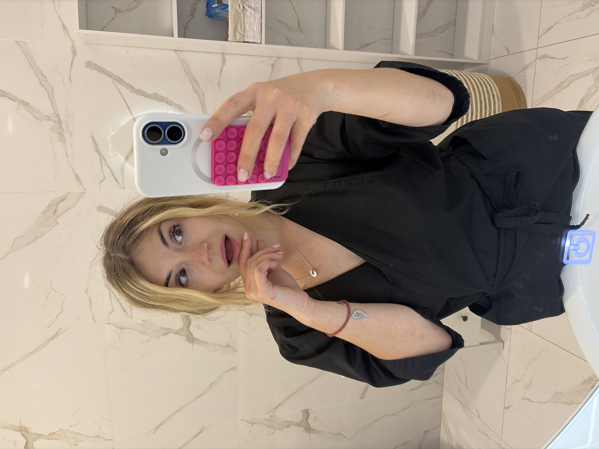A woman is taking a selfie in a bathroom with white marble walls and a white sink. She is wearing a black robe and has a silver necklace and a beaded bracelet with a silver charm. The background includes a shelf unit and a woven basket. There is no information to specify the location in the city of Budva, Montenegro.