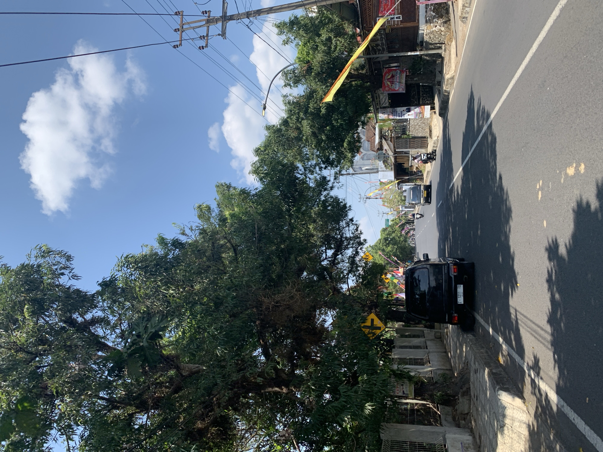 This image depicts a sunny day on a street in Ciamis, Indonesia. The road is paved with asphalt and has a white line marking the lane. Tall trees with lush green foliage line the left side of the street, casting shadows on the road. On the right side, there are buildings, some with stone foundations and tiled roofs. Several flags, some yellow and red, are visible, possibly indicating a local celebration or event.
A black van is parked on the side of the road, facing away from the camera. The license plate is visible. Further down the road, a few motorcycles are in motion, suggesting moderate traffic. Overhead, power lines and poles are present, typical of urban infrastructure. The sky is blue with scattered white clouds. A street lamp is also visible on a pole extending over the road. The overall scene is one of a typical street in a populated area of Ciamis on a bright day.
