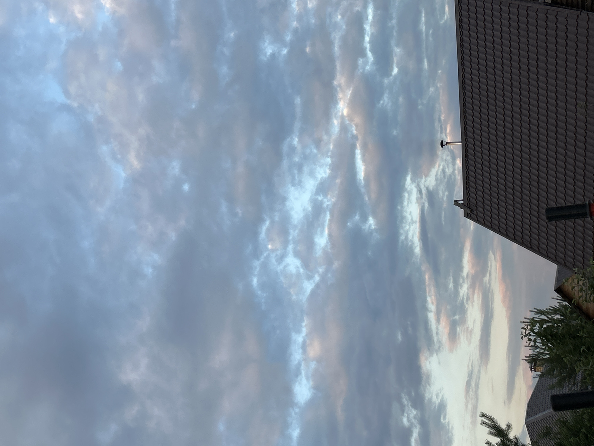 This is a photograph taken outdoors in the city of Chaikovsky, Russia, likely in a suburban or rural setting. The upper portion of the image is dominated by a cloudy sky with varying shades of gray, blue, and hints of pink, suggesting it could be dawn or dusk. The clouds are textured and appear to be moving. In the lower part of the frame, the dark, tiled roof of a house is visible, with a metal chimney extending upwards. To the left and right of the roof, the tops of evergreen trees and foliage can be seen. The overall scene is still and quiet, with no people or animals present.