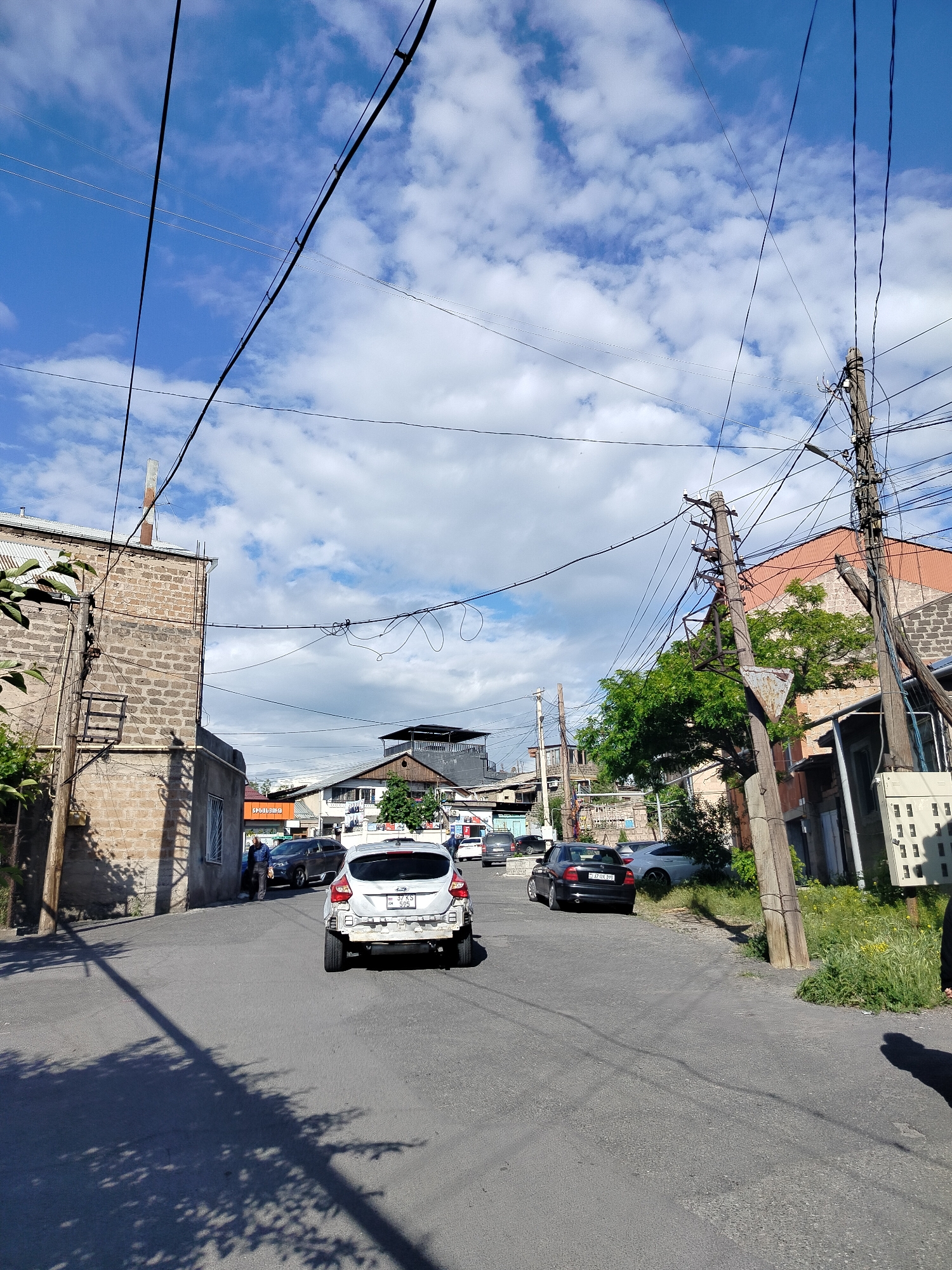 The image depicts a street scene in Yerevan, Armenia. The road is asphalt, with some shadows from trees. A white hatchback car is moving away from the viewer. Several parked cars are visible along the sides of the street. Buildings line both sides of the street, with brick and other materials. Electrical lines are strung across the sky. There are power poles on the right side of the street. Green foliage is present. A person is walking on the left side of the street.