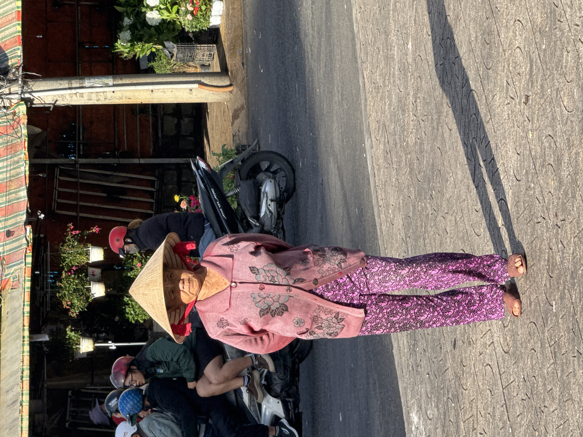 The image captures a street scene in Da Lat, Vietnam, likely during daylight hours given the strong shadows. In the foreground, an elderly woman stands on a textured paved sidewalk, her face partially obscured by a traditional conical hat. She is wearing a pink cardigan with floral embroidery and matching pink patterned pants. She has her hands clasped behind her back. Her feet are clad in brown sandals.
Behind her, a motorcycle is parked. On the motorcycle and to the left, several individuals are seated, some wearing helmets. A woman in a green jacket is visible, and another person in a black jacket.
The background shows a building with an awning, a utility pole, and potted plants displayed for sale. The street is paved with asphalt. The overall atmosphere suggests a typical urban environment with everyday activity.