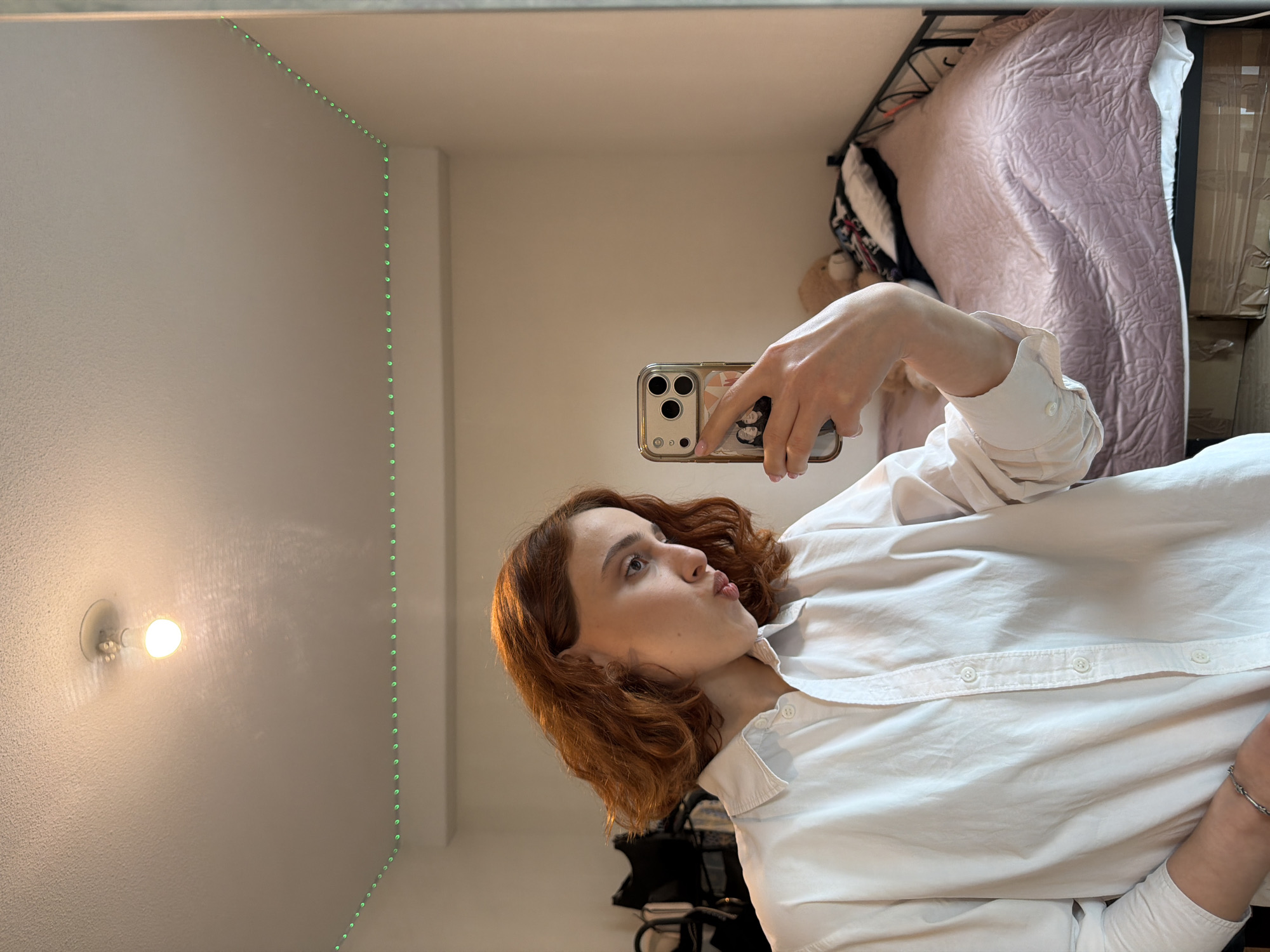 A young woman with short, reddish-brown hair is taking a mirror selfie in a dimly lit room. She is wearing a white button-down shirt and has a pouting expression. Her phone, with a gold case and a few small photos attached, is visible in her hand. In the background, a bed with a pink comforter and pillows, a teddy bear, and a black metal bed frame are present. The room has plain walls and a white ceiling with a single light fixture. Green LED strip lights are visible along the top edge of the mirror. The scene suggests a bedroom or a similar personal space. The image does not provide any information about a location in Strasbourg, France.