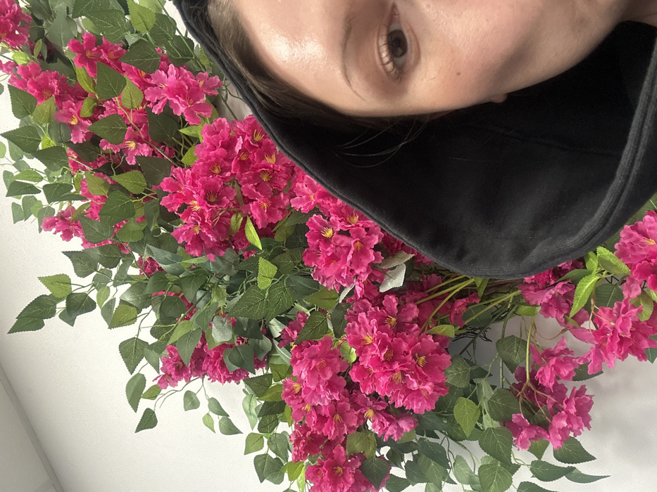 A person wearing a black hooded top is positioned behind a vibrant display of fuchsia flowers and green foliage. The camera angle is from below, looking up at the person, who appears to be slightly off-center. Their face is partially visible, with one eye and some forehead showing. The flowers create a dense, visually striking frame around the person.