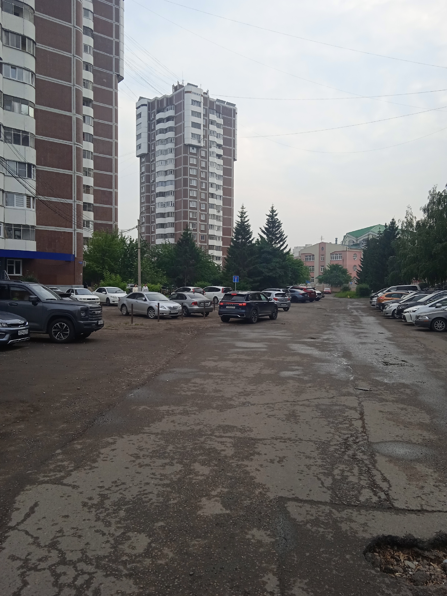 This is a street-level view of a parking lot in the city of Krasnoyarsk, Russia. The parking lot is asphalt and appears to be in poor condition with numerous potholes and cracks. Several cars are parked in the lot, including a dark gray SUV in the foreground, a silver sedan, and a dark blue SUV in the distance. In the background, there are several apartment buildings of varying heights, trees, and a cloudy sky. Two people are standing near the silver sedan. The overall scene depicts a residential area with a functional, albeit worn, parking facility.