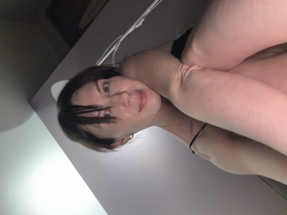 A low-angle, upward-facing image captures an adult female subject in an interior setting. She has light skin, dark hair with bangs, and light-colored eyes, looking directly at the camera with a subtle smile. She is partially clothed, with a thin black strap visible on her shoulder, indicative of a top or undergarment. Her posture suggests she is seated or reclining, with her knees drawn upward, and her left arm (viewer's right) is positioned across or resting on her bent knees. The background features plain, light-colored walls or surfaces, appearing light grey or faint lavender. A bright, unidentifiable light source is prominent in the upper left corner, casting light into the scene. A thin white cable or wire is faintly visible extending downwards on the right side of the frame. The specific location within the city of Perm, Russia, cannot be determined from the visual information present in the image.