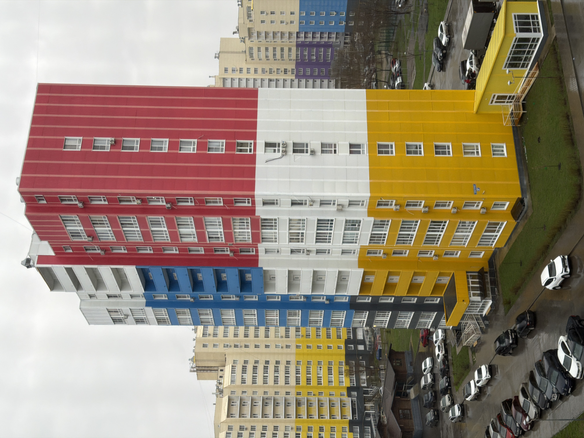 The image shows a multi-story building in Засечное, Russia, painted in alternating horizontal stripes of red, white, yellow, blue, beige, and gray. The building has a rectangular shape and is covered in windows. In the foreground, there is a parking lot with cars parked in a circle. To the right of the building, there is a grassy area. The sky is overcast.