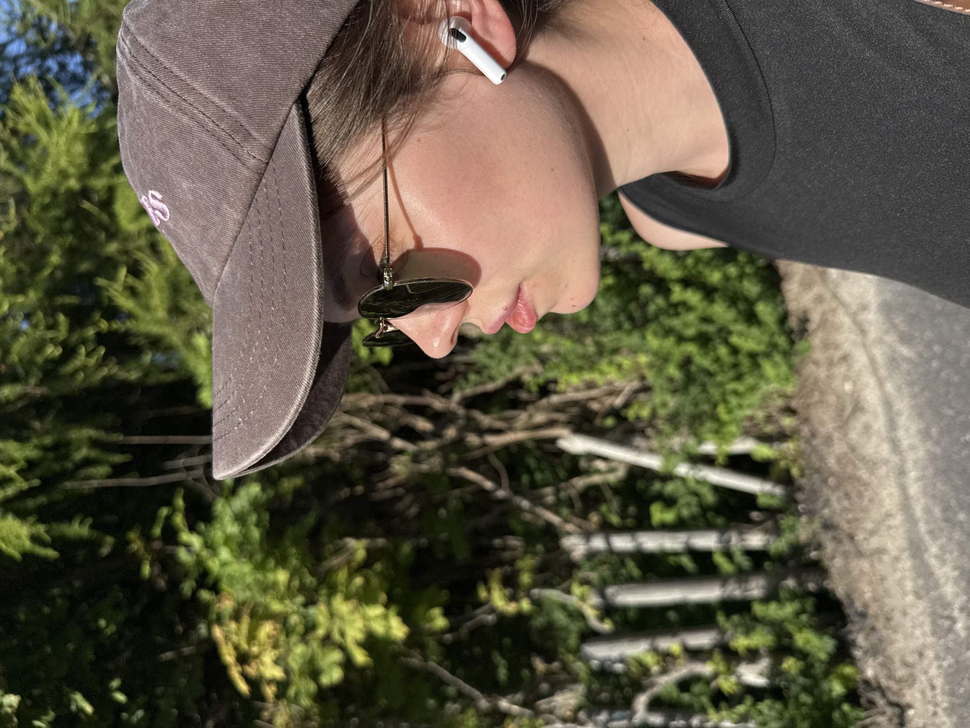 A medium shot photograph captures a young person from the side, wearing a brown baseball cap, sunglasses, and a black tank top. They have a white wireless earbud in their right ear. The background consists of lush green trees and a wooden fence. The setting appears to be outdoors, likely in a park or natural area. The lighting suggests it is daytime, with sunlight casting shadows. There is no indication of the specific location within Szklarska Poręba, Poland.