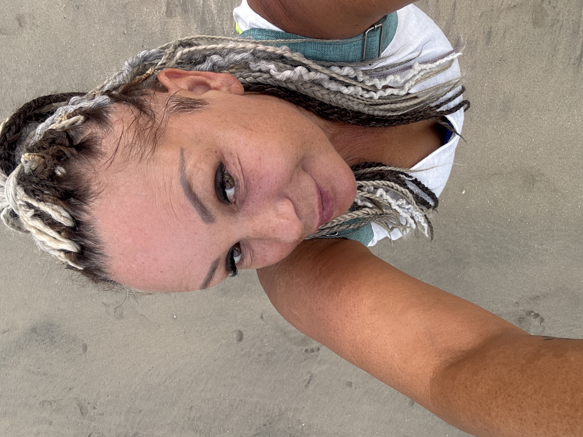 A woman poses for a close-up selfie on a sandy beach. Her head is tilted, with her face toward the camera. She has braided hair in a mix of dark brown and blonde colors. Her skin tone appears to be tanned, and she wears a white shirt with a light blue fabric band around her neck. The woman's left arm is visible, extending from her body towards the bottom of the frame, suggesting she is holding the camera. The background is a flat, sandy surface, typical of a beach. Given the prompt's details, it is plausible that the location is Jimbaran, Indonesia.