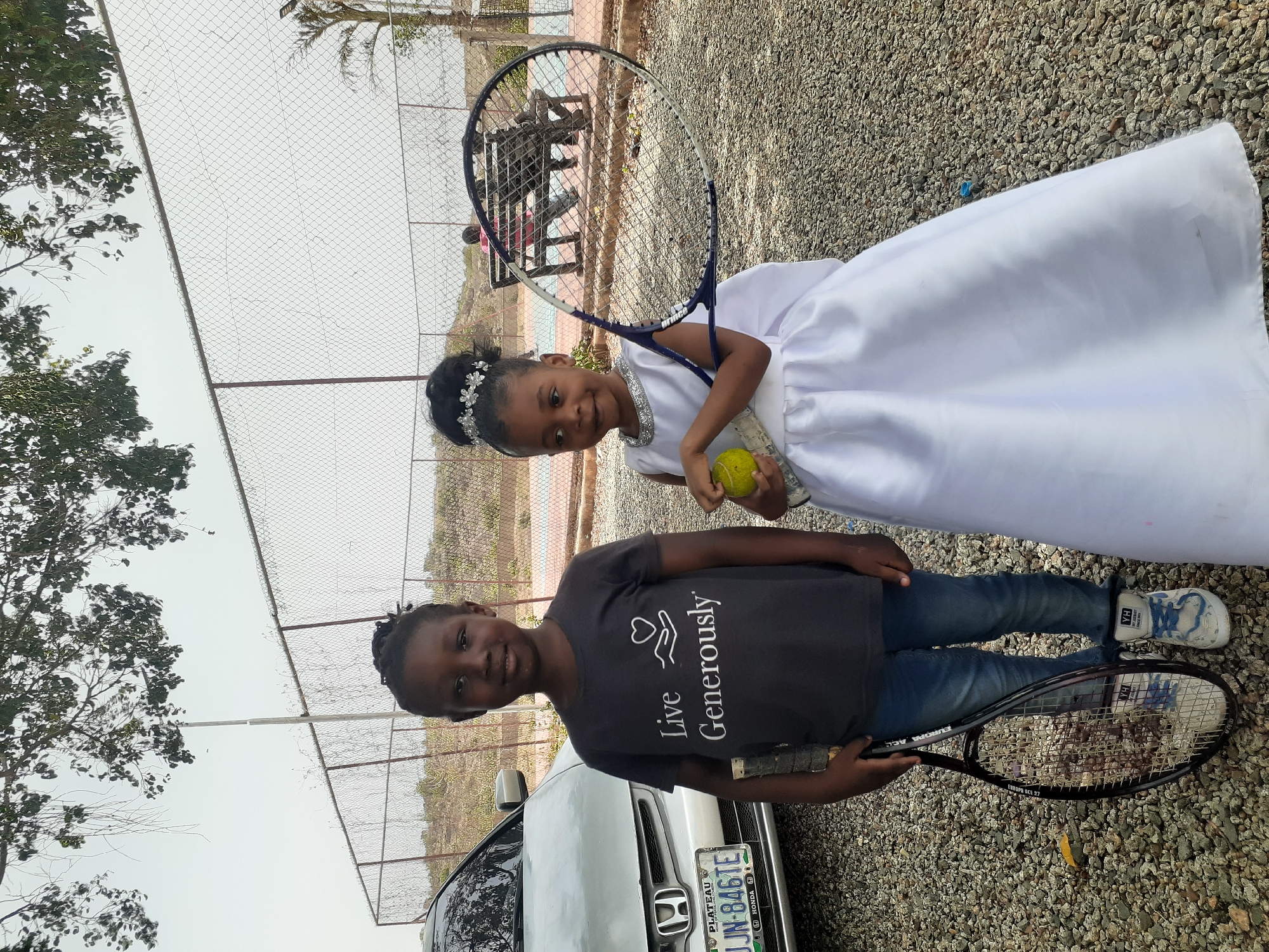 Two young girls are posing outdoors with tennis equipment on a gravel surface, with a chain-link fence in the background. The image is bright, suggesting daytime with an overcast sky.
The girl on the left, who appears slightly older, has dark skin and her dark hair is styled in small braids or dreadlocks. She wears a dark grey t-shirt with the text "Live Generously" and a heart-and-hands icon printed in white, blue jeans, and white-and-blue sneakers. She holds a black tennis racket, with "EUROPA ACE 27" visible on its frame. Behind her, a silver Honda car is partially visible, displaying a Nigerian license plate from Plateau State, reading "JUN-846TE".
The younger girl on the right has dark skin and dark hair styled with a sparkling, tiara-like headband. She is dressed in a white, sleeveless formal dress adorned with silver beaded detailing around the neckline. She holds a tennis racket in her left hand and a yellow tennis ball in her right, looking towards the camera with a bright smile.
Behind both girls is a tall chain-link fence, through which hints of a blue-surfaced area, possibly a tennis court, are visible, along with distant foliage and trees on the far left. Both girls appear happy and are posing for the camera.