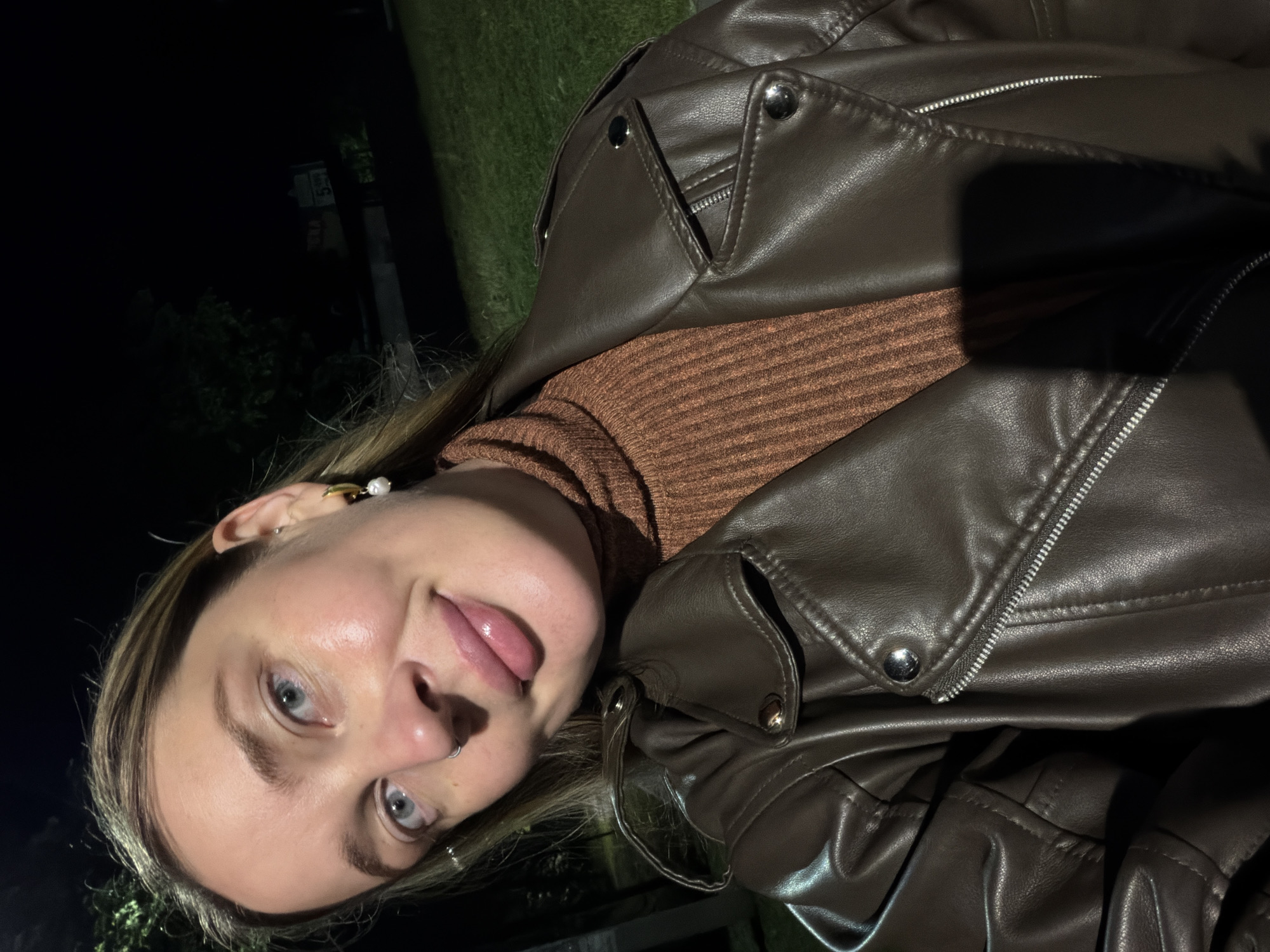 The image is a close-up, portrait-oriented selfie taken at night, featuring a young person, likely female. The subject, looking directly at the camera, has blue-grey eyes, and is playfully sticking their tongue out. They wear a septum piercing and a pearl-and-gold-colored earring on their right ear.
The person is dressed in a dark brown faux leather jacket, featuring prominent zippers and snap buttons, over a ribbed, rust-brown turtleneck or sweater.
The background is very dark, indicative of nighttime, with minimal details discernable. Faint outlines of green foliage, possibly trees or bushes, and some ground cover are visible. In the deep background, indistinct shapes suggest urban elements like poles or structures. There are faint, blurry white characters visible on one such distant element, possibly part of a sign, with one segment resembling "53" and another less clear, possibly "RA" or "KA". The subject is well-illuminated, likely by a flash or direct artificial light, contrasting sharply with the dark surroundings. The scene is set in Chaikovsky, Russia, according to the provided context.