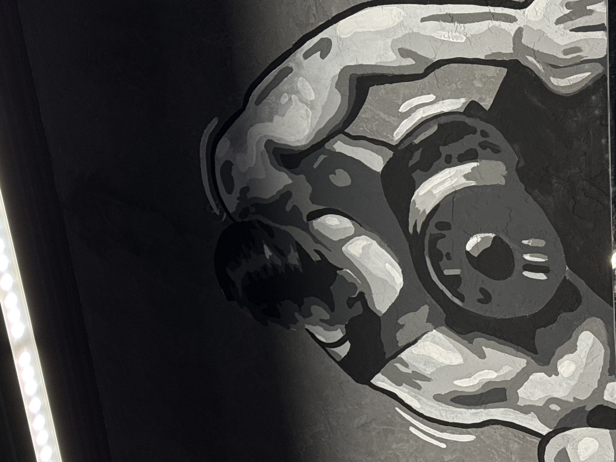 The image depicts a grayscale mural or artwork featuring a stylized, cartoonish figure holding a device to its face. The figure appears to be wearing some kind of headgear or helmet. The mural is situated on a wall, likely indoors, as suggested by the artificial lighting on the left edge of the frame. There are no other people or recognizable objects in the scene. There is no information available to indicate that this image is located in Votkinsk, Russia.