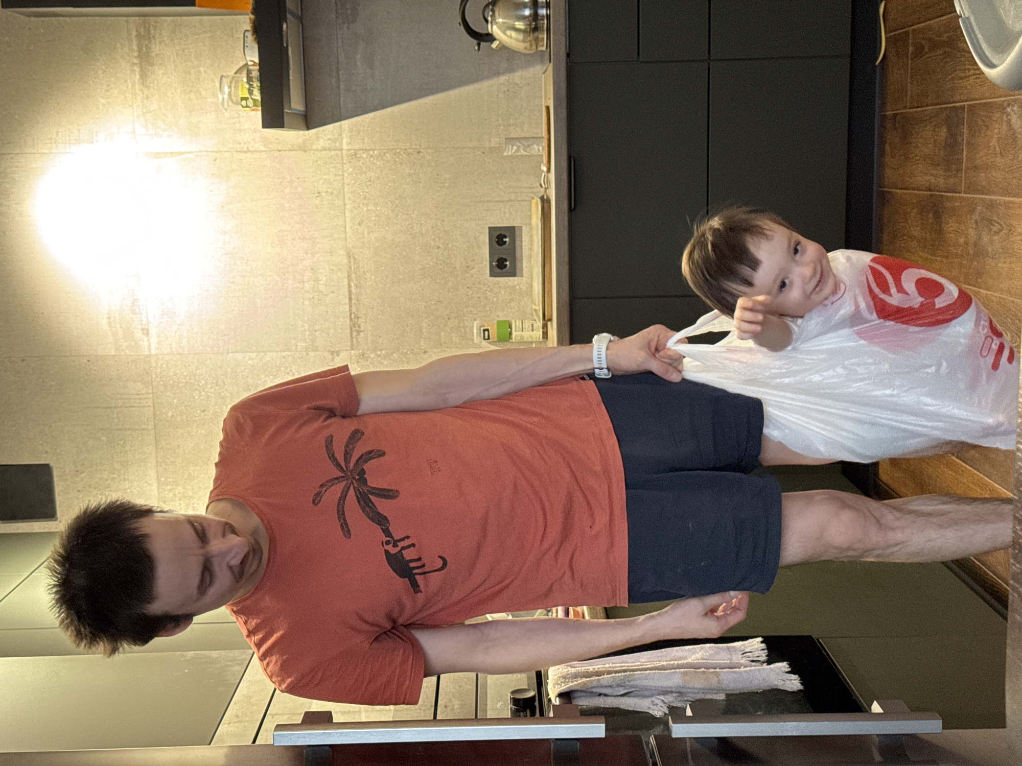 The image shows a man standing in a kitchen with a young child inside a plastic bag. The man is wearing an orange t-shirt with a palm tree and elephant design, navy blue shorts, and a white fitness tracker on his wrist. He is holding the opening of the plastic bag. The child is inside the bag and is smiling at the camera. The kitchen has light-colored tile walls, dark cabinets, wood-look flooring, and a stainless steel kettle is visible on the counter. It is not possible to determine if this scene is taking place in the city of Izhevsk, Russia.