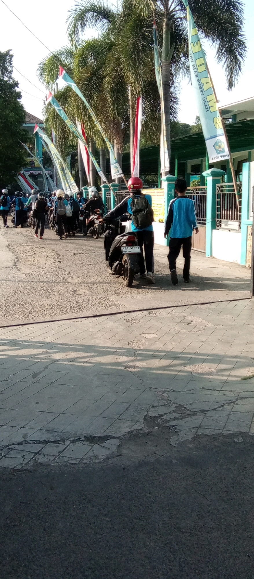 In Kediri City, Indonesia, a group of students, some on motorcycles, are seen at what appears to be the entrance of a school. The scene is outdoors with palm trees and flags lining the pathway. The students are wearing uniforms, with some on motorcycles and others walking. One student in the foreground is on a motorcycle, facing away from the camera, wearing a red helmet and a blue shirt with black sleeves. Another student in a blue long-sleeved shirt walks away from the motorcycle towards the right of the frame. The ground is paved with a rough texture and some cracked tiles. A green fence is visible in the background, suggesting the boundary of the school grounds. The sky is clear, indicating a bright, sunny day.