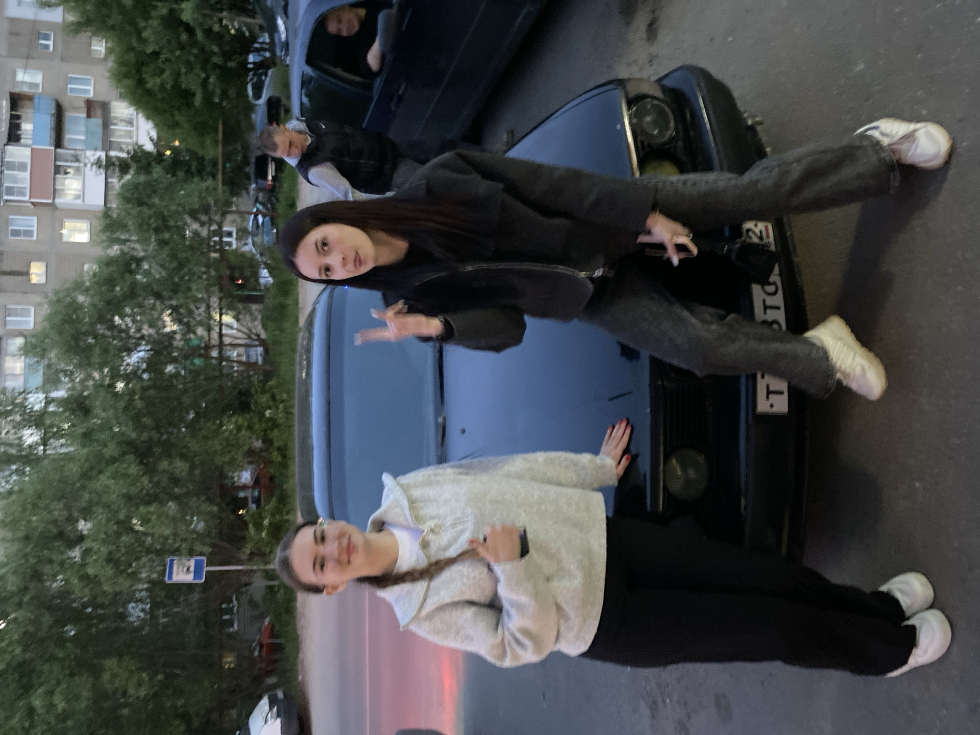 The image depicts a casual outdoor scene at dusk or evening, featuring four individuals and a dark-colored car. In the foreground, two young women are posing around the front of the car, which is parked on asphalt.
One woman is sitting on the car's hood, facing the viewer with a subtle smile. She wears a dark jacket, dark jeans, and white sneakers, making a peace sign with her right hand while holding a phone in her left. Her long dark hair is loose. To her left, another young woman leans against the car's front bumper, also looking towards the camera with a gentle smile. She is dressed in a light-colored, possibly textured, jacket or sweatshirt, dark pants, and white sneakers. Her hands, with visible red nail polish, rest on the car's grille, and a long braid is draped over her right shoulder.
The car is dark, appearing black or dark blue, with visible headlights. A partial license plate is on the front bumper, displaying the characters "T3TO". In the background, to the left, are multi-story residential apartment buildings with some windows illuminated, confirming the evening time. Green trees are prominent between the buildings and the street. Two other individuals are visible further back; one appears to be inside a vehicle, and another stands nearby. A blue and white road sign, possibly a bus stop marker, is also present among the trees. The overall atmosphere is relaxed and informal.