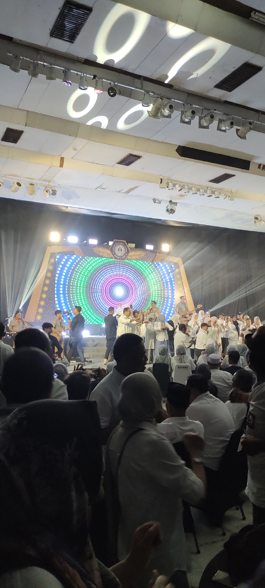 An indoor event is taking place, likely a concert or religious gathering, in Lhokseumawe, Indonesia. A large LED screen serves as the backdrop for a group of performers on a stage. The screen displays a colorful, circular pattern resembling a ripple or a target. Several individuals, dressed in white, are on stage, some appearing to play musical instruments like guitars, while others are clapping or gesturing. The audience, also predominantly in white attire, is seated in front of the stage, with some individuals standing. The ceiling features lighting fixtures and projected patterns, indicating a staged event. The overall atmosphere seems celebratory and engaging.