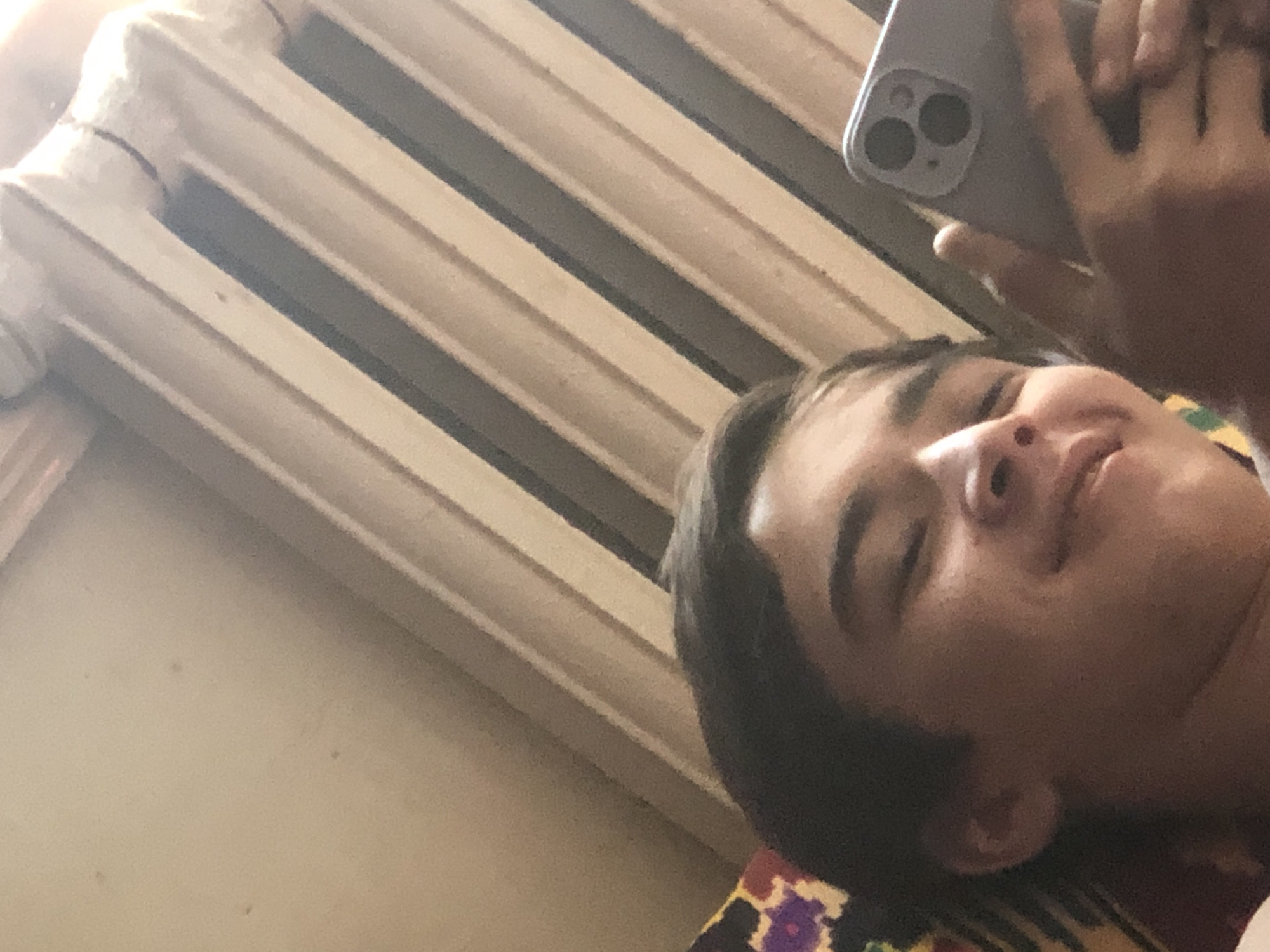 A person is lying down and taking a selfie with their phone. The person is a young male with dark hair. He is holding a light-colored smartphone with a triple-camera system in his right hand. The background appears to be a wall with a radiator. The image is taken from a low angle, looking up at the person. The person is smiling slightly.