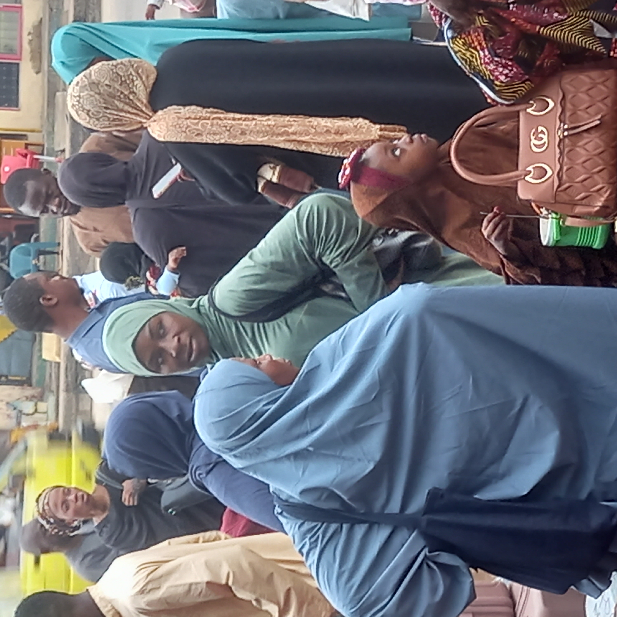 The scene depicts a group of people, mostly women, gathered outdoors in Dadi Riba, Nigeria. The majority of the women are wearing hijabs of various colors, including blue, green, black, and brown. Some of them appear to be looking in different directions. A brown handbag with a visible logo is held by one of the women. A man in a blue shirt is visible towards the left side of the frame, appearing to hold a baby. The environment appears to be an open, public area with a building in the background.