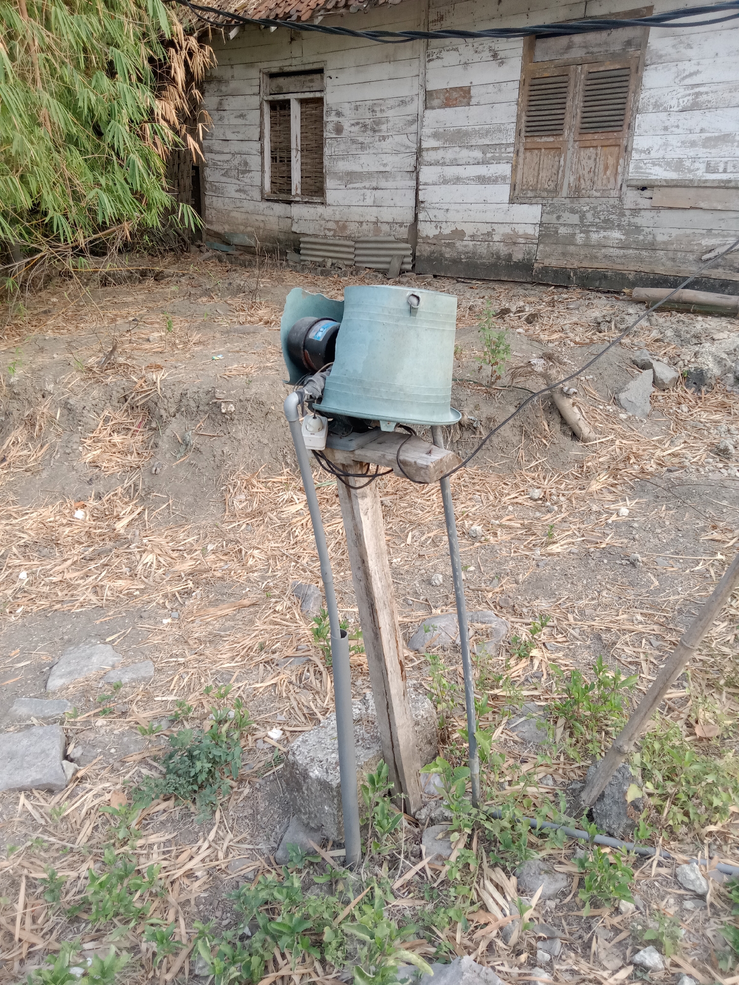 A vintage, weathered building with white, peeling wooden siding and a tiled roof stands in the background. A broken window with visible interior materials and a shuttered window with horizontal slats are present. In the foreground, a green, bell-shaped device, possibly an old horn or siren, is mounted on a wooden post supported by grey pipes and a concrete block. The ground is covered in dry leaves and scattered rocks, with some green vegetation growing around the base of the structure. A power line runs overhead and across the scene. The overall impression is one of neglect and a rural or semi-rural setting, possibly in Pacing, Indonesia.