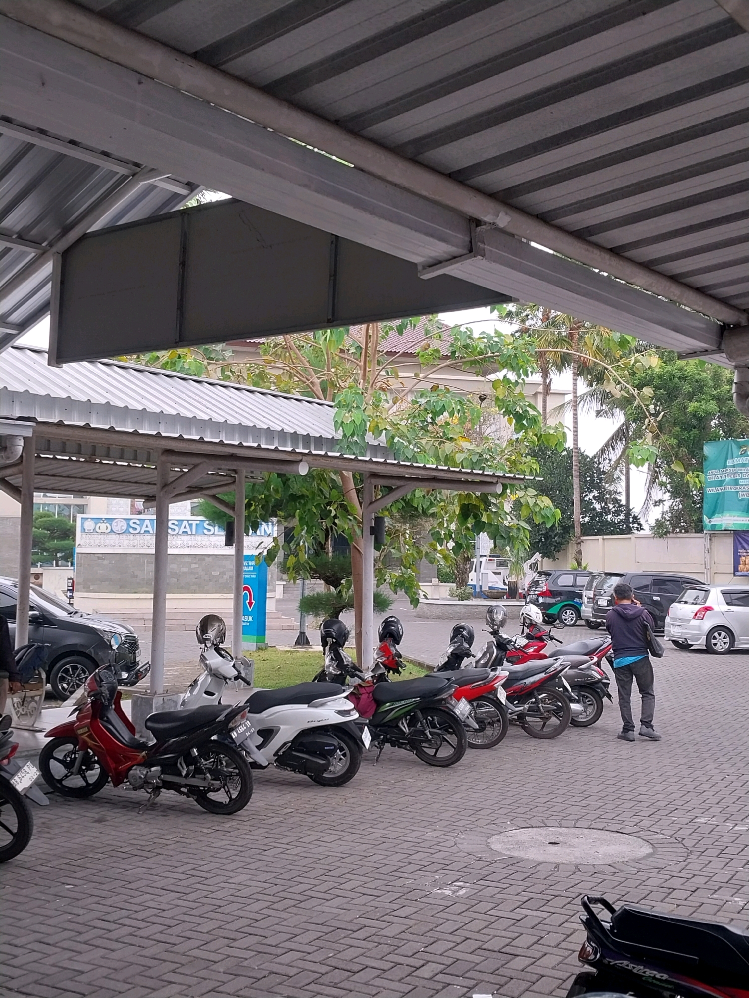 This image depicts a parking area under a covered structure, likely a carport or outdoor shelter, in Sleman, Indonesia. The ground is paved with interlocking bricks. Several motorcycles are parked in neat rows. A black car is visible to the left, and a dark grey car and a silver car are parked further back on the right. A person, seen from behind, is standing to the right of the frame, looking towards the cars. In the background, there are buildings and lush green trees, suggesting an urban or semi-urban environment. One building in the background has a prominent sign with Indonesian text. A tall, thin palm tree is also visible. The overall lighting suggests it is daytime, possibly overcast.
