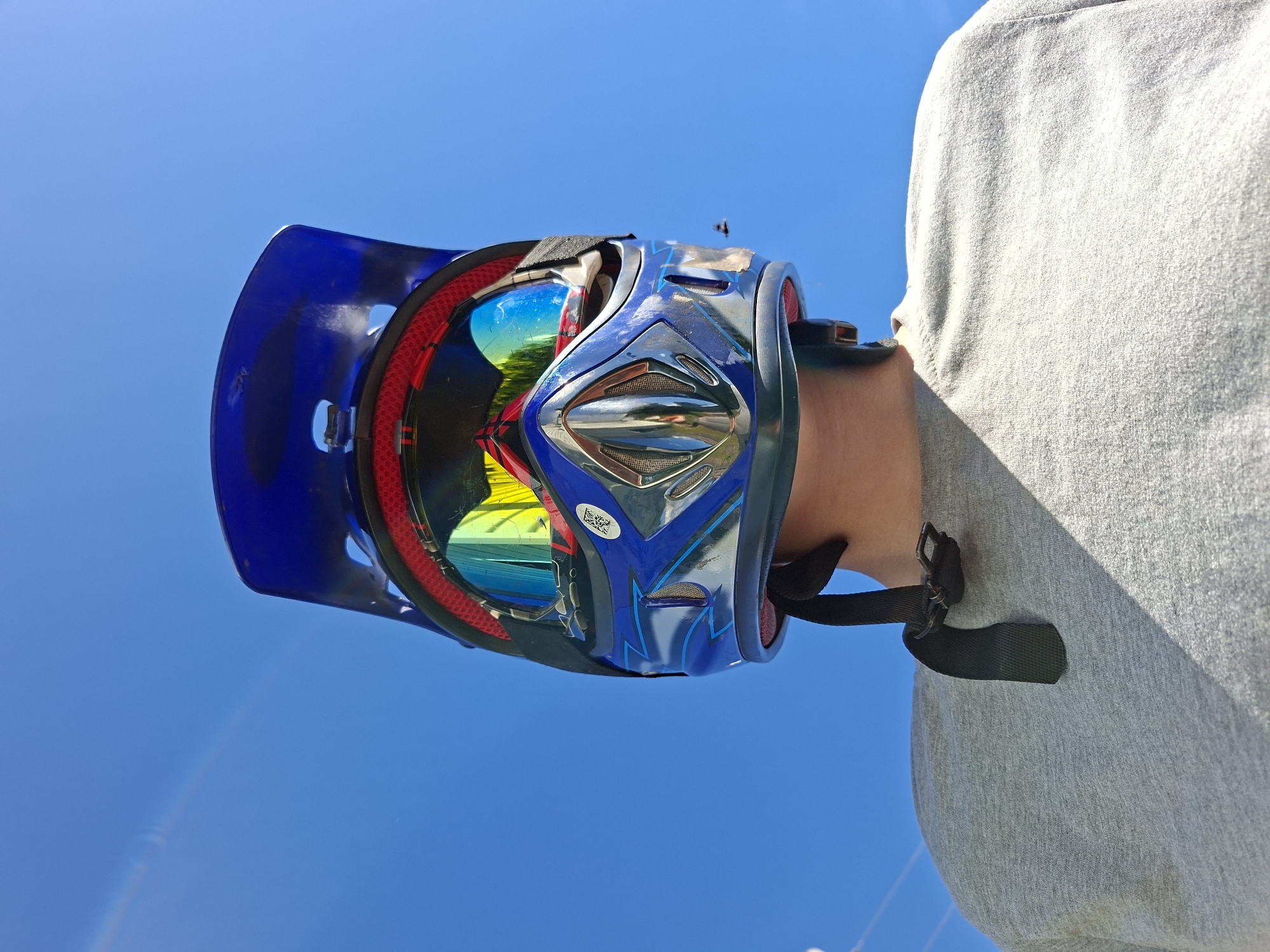 A person wearing a blue motocross helmet with reflective goggles is facing away from the camera, looking up at the sky. The helmet has red trim and a shiny chrome faceplate. The person is wearing a grey t-shirt. The background is a clear blue sky with a faint rainbow streak on the left. The scene appears to be outdoors. There is a small insect flying in the middle right of the image. The image is a low-angle shot, looking up at the subject.
