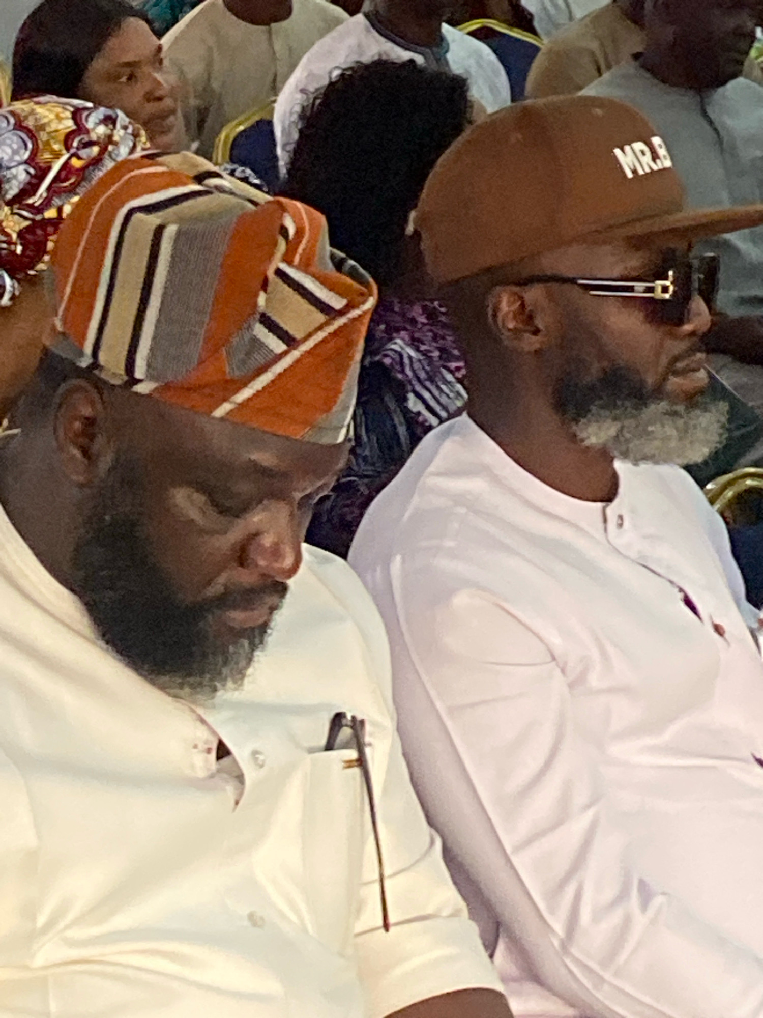 Two men are seated side-by-side, facing the viewer's left. The man on the left wears a white dashiki and a traditional striped headwrap in orange, beige, and grey. He has a dark beard and is looking downwards. A pair of glasses is tucked into the left breast pocket of his dashiki. The man on the right wears a white dashiki and a brown baseball cap with "MR.B" written on it. He has a salt-and-pepper beard and is wearing dark sunglasses. The background shows other people and what appears to be an indoor event or gathering. The image is a close-up shot, with the focus on the two men.