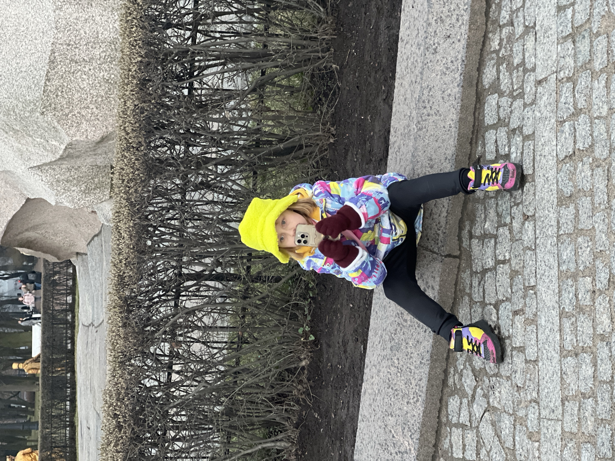 A young child, dressed in a yellow hat, a colorful winter jacket, black leggings, and vibrantly colored sneakers (pink, yellow, black, purple), is seated on a stone ledge, holding a smartphone and taking a picture. The location is near a monument or sculpture constructed from rough-hewn stone, situated within a park or public space. Behind the child is a line of dense, low-lying bushes with bare branches. The ground surrounding the bushes appears to be dirt or mulch. A stone paved pathway extends from the ledge. In the background, a glimpse of other people can be seen, and there is a tall fence. This photograph was taken near the Bronze Horseman statue on Senate Square, Saint Petersburg, Russia.