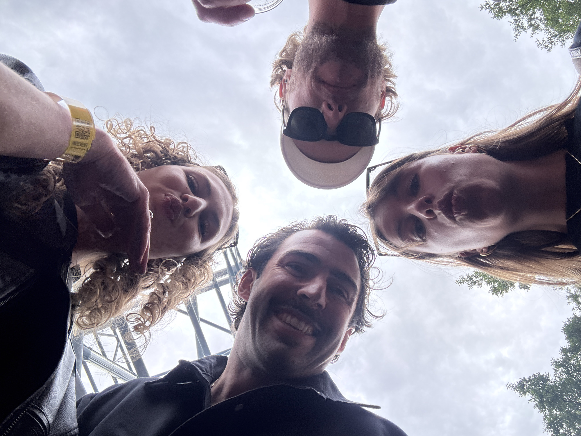 This is a first-person perspective, low-angle selfie taken in Zaandam, Netherlands. Four individuals are looking down at the camera. From left to right, the first person has dark hair and a beard, and is smiling with his teeth showing. The second person has curly brown hair and is looking towards the camera. The third person is wearing sunglasses and a white cap, and has a beard and blonde hair. The fourth person, in the foreground, has long brown hair and is making a puckering expression with her lips. The background shows a cloudy sky with some trees visible. The individuals appear to be outdoors, possibly at an event given the wristband visible on the arm of the second person. The location within Zaandam is not specified.