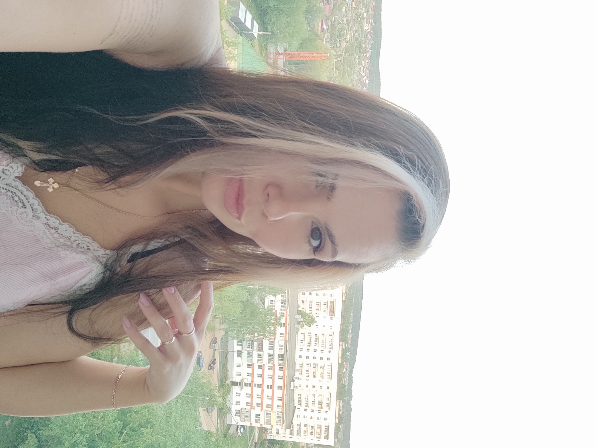 A woman is posing for a selfie. She has dark hair with a bleached streak near her face. She is wearing a pale pink top with lace trim. A gold necklace with a cross pendant is visible. She has a gold bracelet and rings on her left hand. Behind her, a cityscape can be seen, including apartment buildings and green spaces. A construction crane is visible in the distance. Based on visual cues, the location is potentially in or near Chaikovsky, Russia.